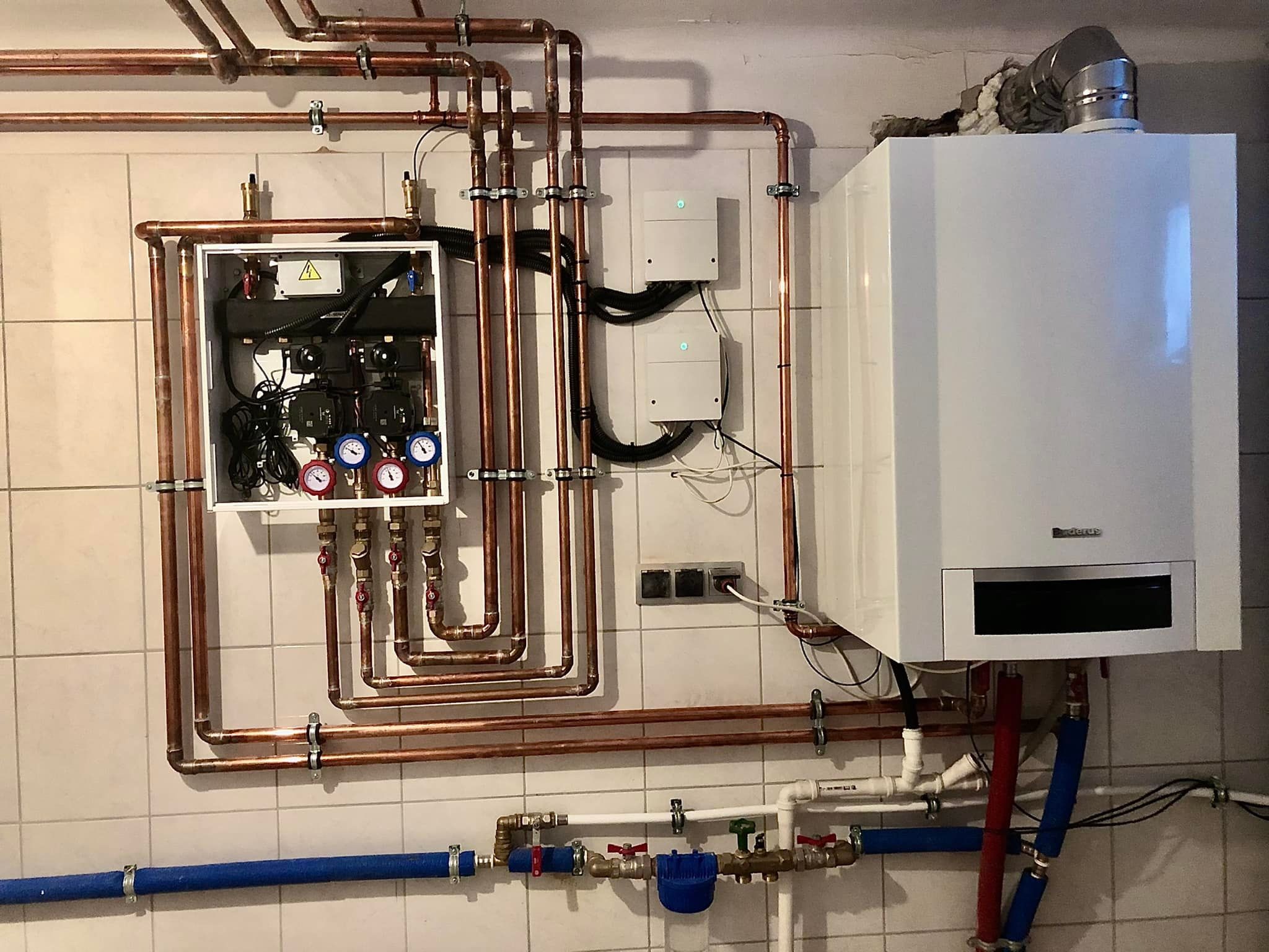 Złożona instalacja grzewcza z miedzianymi rurami, białą skrzynką rozdzielczą z oznaczeniem ostrzegawczym, kotłem gazowym Buderus i dwoma białymi modułami sterującymi na kafelkowanej ścianie.