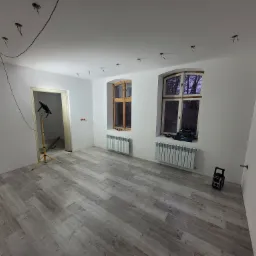 Pokój po remoncie z panelami podłogowymi w odcieniach szarości, świeżo pomalowanymi ścianami na biało, dwoma oknami z drewnianą stolarką, grzejnikami pod oknami, lampą budowlaną na statywie...