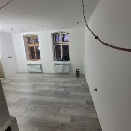 Pokój po remoncie: świeżo ułożone panele podłogowe, białe ściany z dwoma oknami, nowe grzejniki pod oknami, widoczne przewody elektryczne na suficie, narzędzia na parapecie.