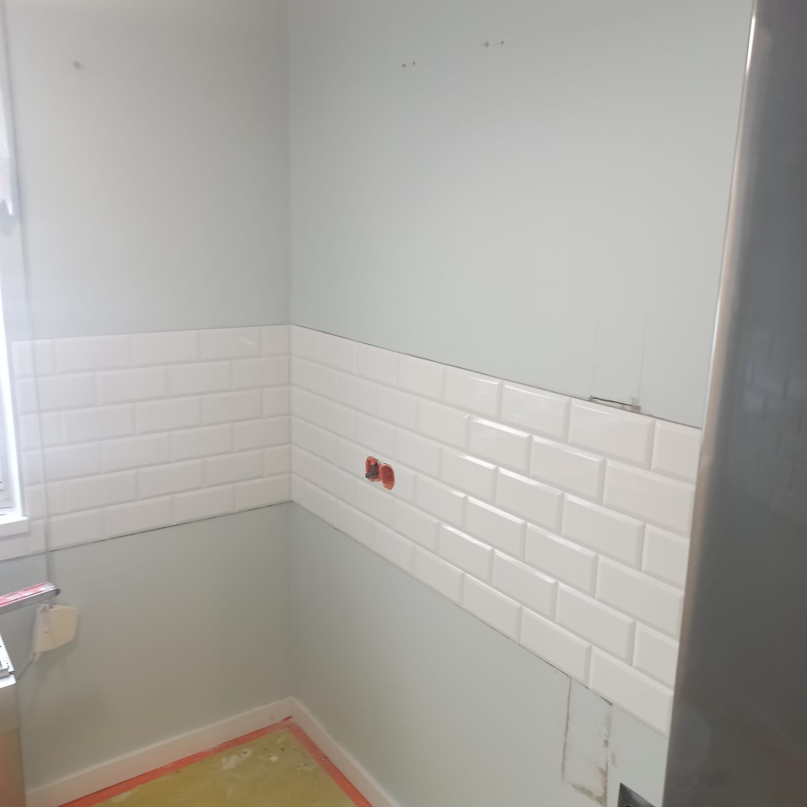 Świeżo ułożone białe, błyszczące płytki typu metro na ścianie w trakcie remontu kuchni, widoczne puszki elektryczne i fragment podłogi zabezpieczonej folią.