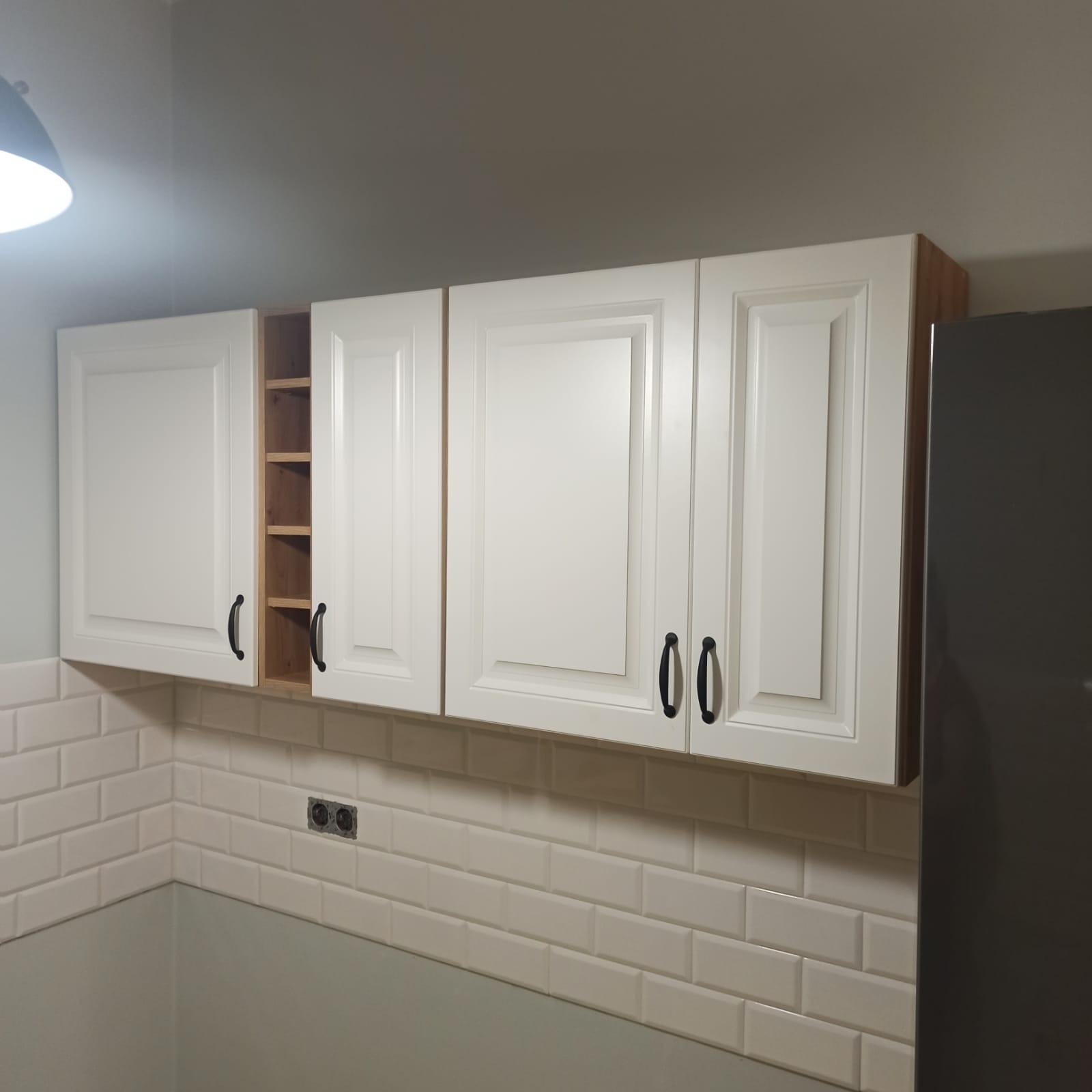 Białe, wiszące szafki kuchenne z czarnymi uchwytami, jedna szafka otwarta, odsłaniająca półki. Widoczna ściana z białych płytek typu metro i fragment szarej lodówki.