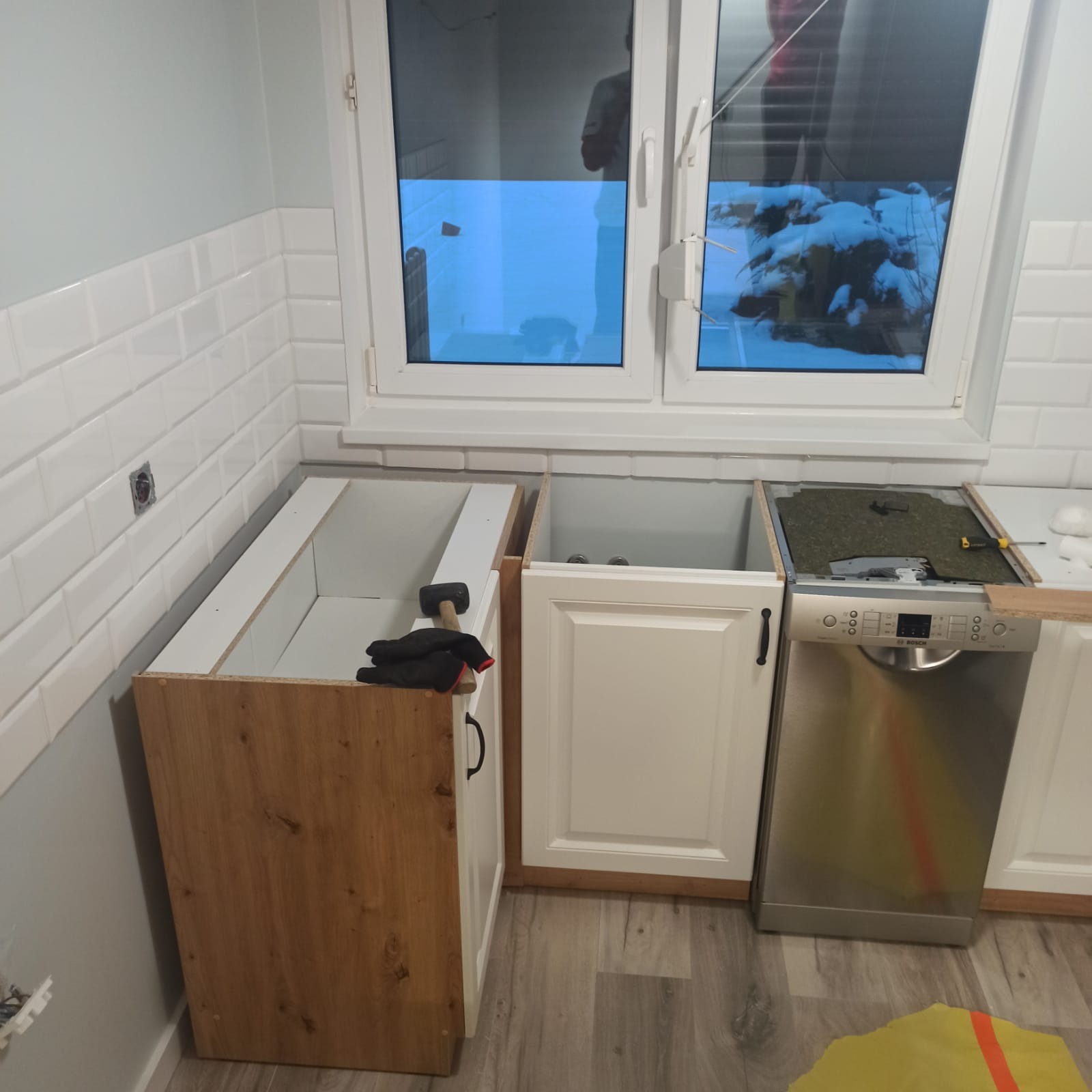 Montaż szafek kuchennych: surowa drewniana podstawa, białe fronty, widoczna zmywarka Bosch, okno z widokiem na ośnieżone drzewa.