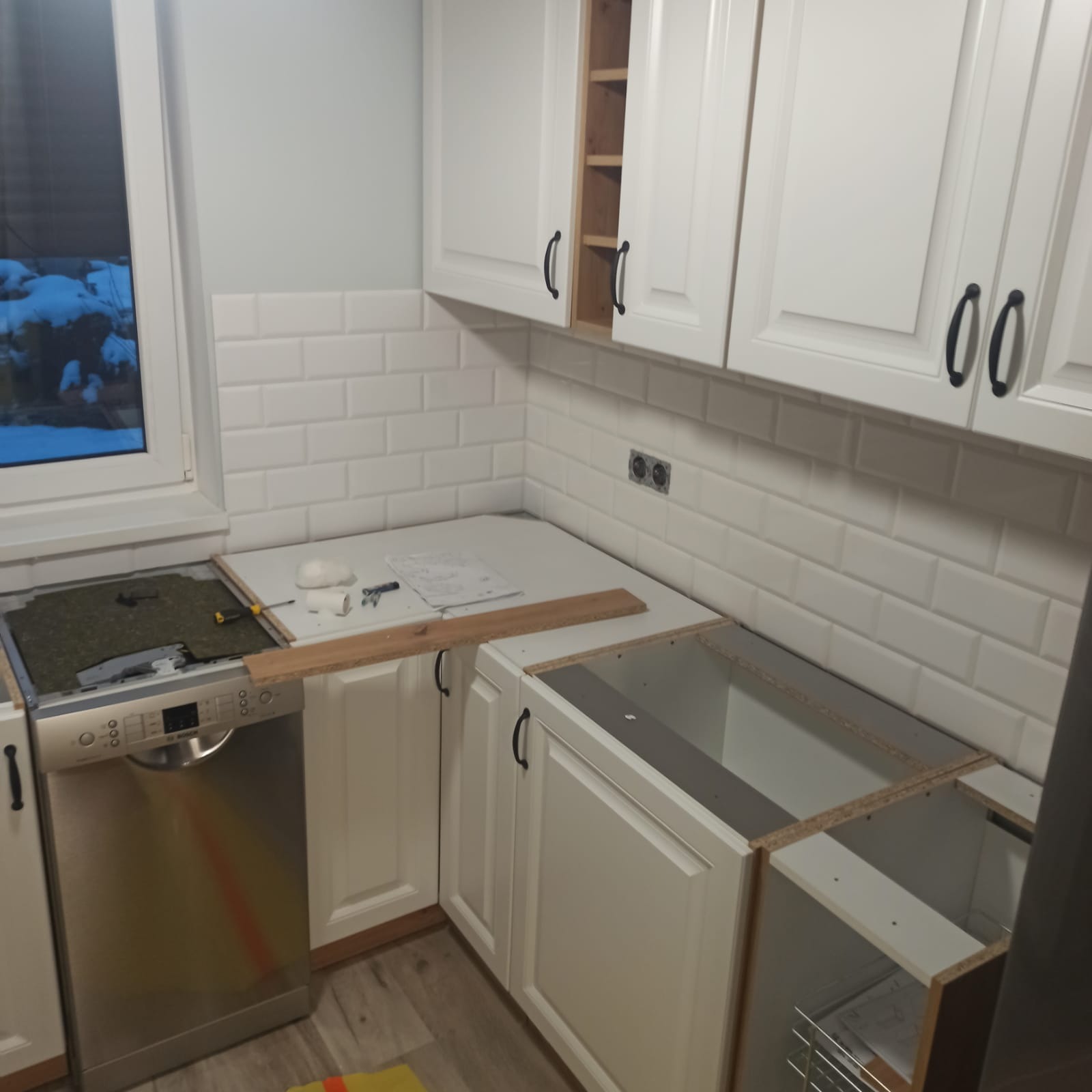 Montaż białych szafek kuchennych z czarnymi uchwytami, widoczna zabudowa zmywarki i fragment blatu, na blacie leżą narzędzia i dokumentacja, w tle okno z widokiem na ośnieżone drzewa.