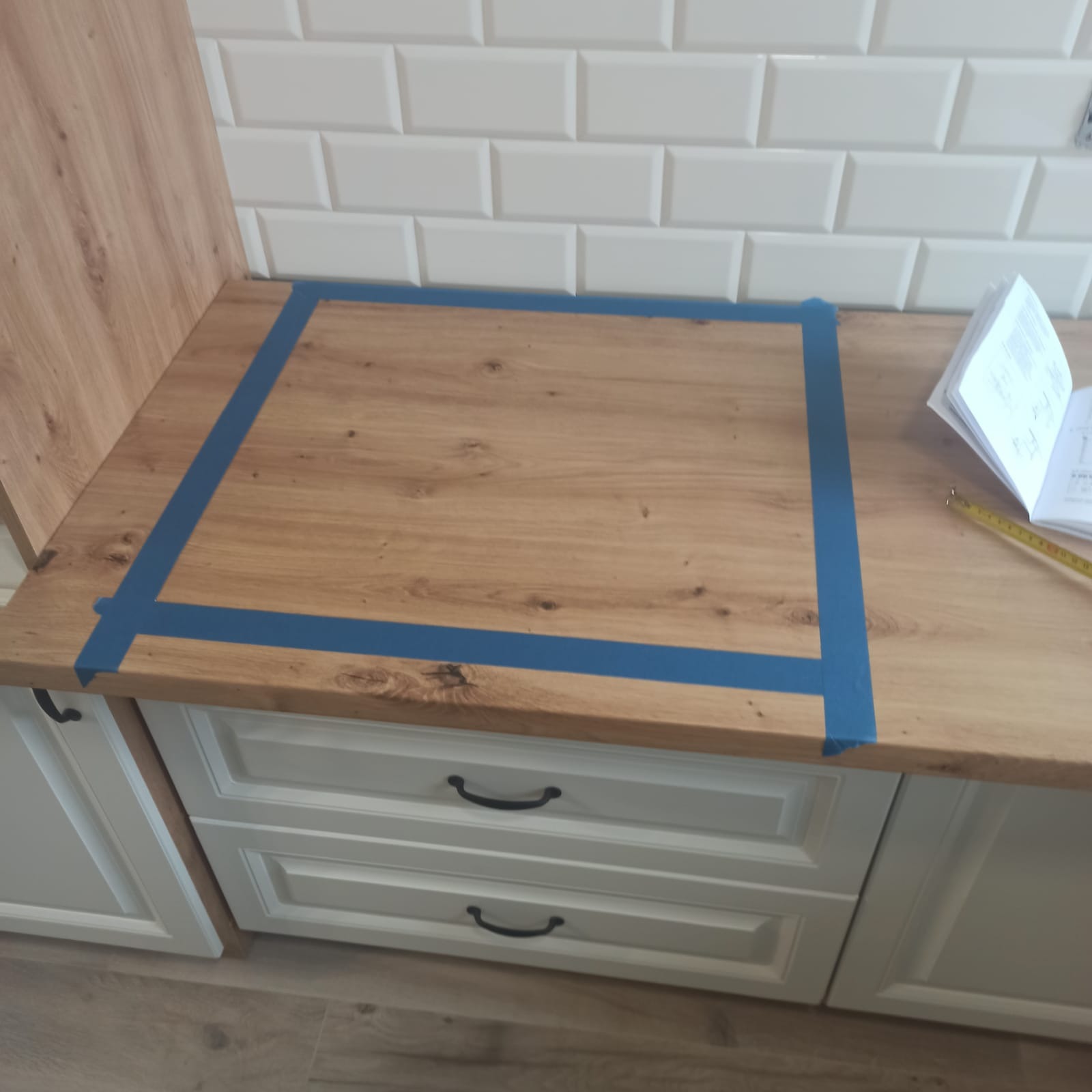 Przygotowanie blatu drewnopodobnego w kuchni w Jastrowiu, wyznaczone granice niebieską taśmą malarską przed montażem płyty grzewczej, widoczne białe płytki na ścianie i instrukcja montażu.