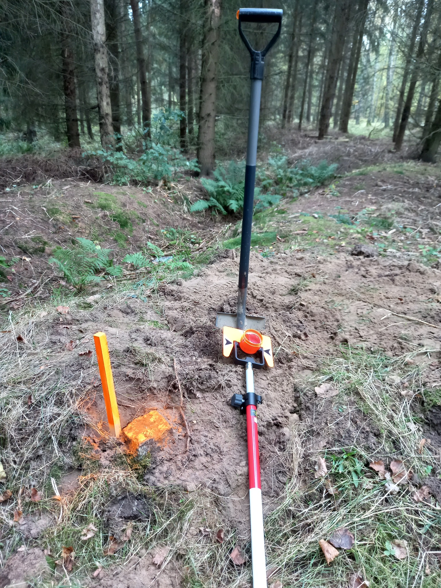 Pomarańczowy palik wbity w ziemię, obok tyczka geodezyjna z pryzmatem, łopata wbita w ziemię, teren leśny.