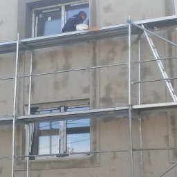Edward Gryniewski - Pracownik na rusztowaniu aplikuje masę wyrównującą wokół okna na elewacji budynku, widoczny kubeł z materiałem.