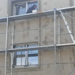 Pracownik na rusztowaniu aplikuje masę wyrównującą wokół okna na elewacji budynku, widoczny kubeł z materiałem.