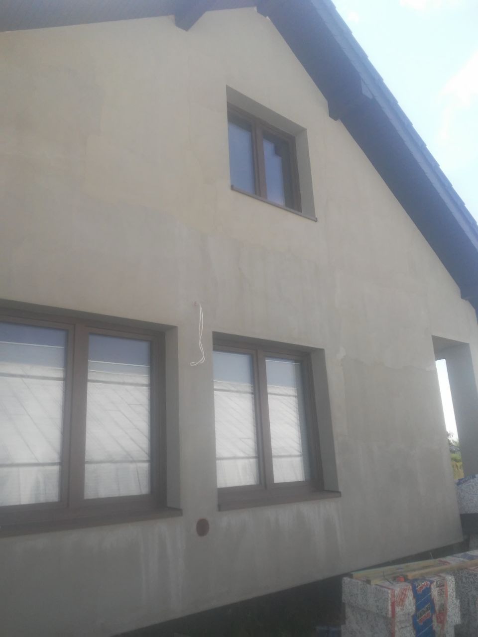 Dom jednorodzinny z nałożoną warstwą szpachli na elewacji, przygotowany do dalszych prac wykończeniowych, widoczne brązowe ramy okienne i materiały budowlane u podstawy ściany.