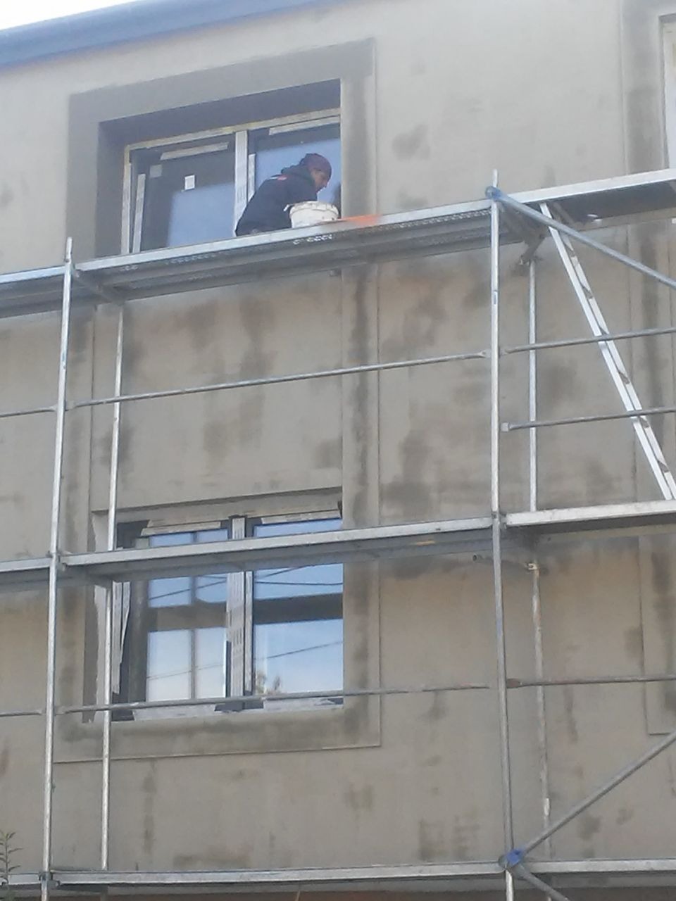 Pracownik na rusztowaniu aplikuje masę wyrównującą wokół okna na elewacji budynku, widoczny kubeł z materiałem.