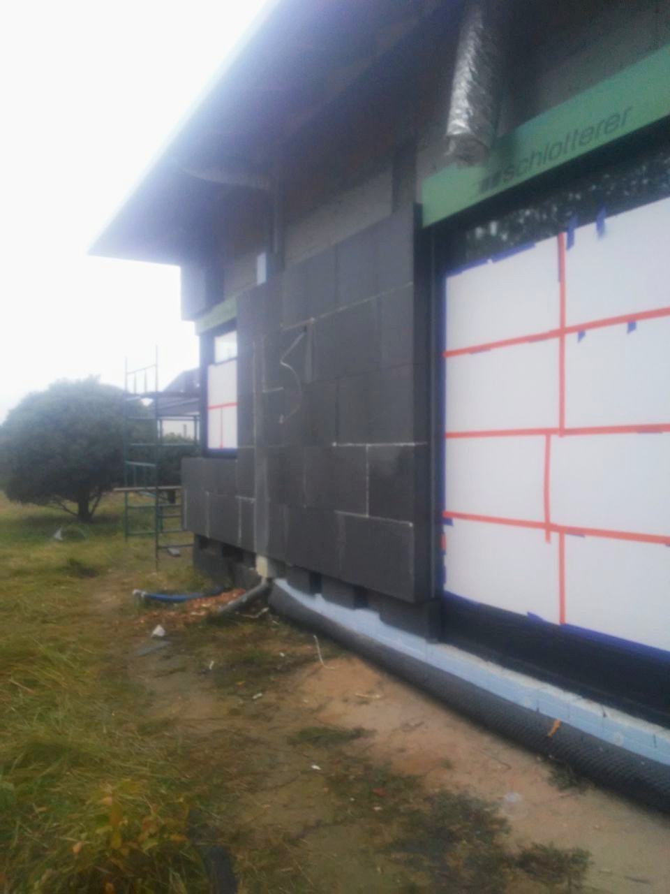 Częściowo ocieplona ściana zewnętrzna budynku z widocznymi panelami izolacyjnymi z ciemnego materiału, zabezpieczonymi oknami i fundamentami, na tle trawnika i drzewa.