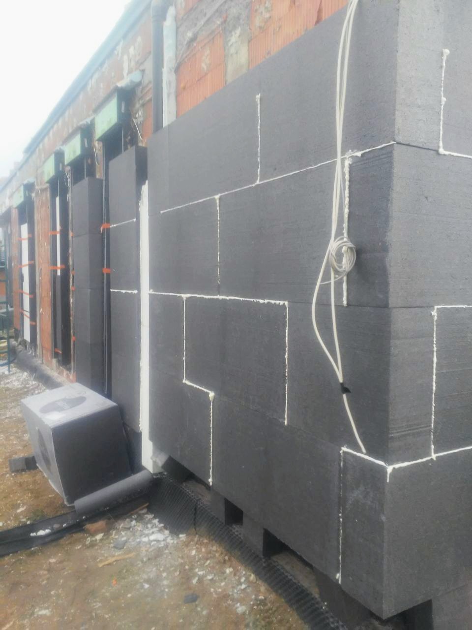 Montaż ciemnoszarej izolacji termicznej na ścianie budynku, widoczne białe spoiny i wystające kable, fragment elewacji w trakcie prac.