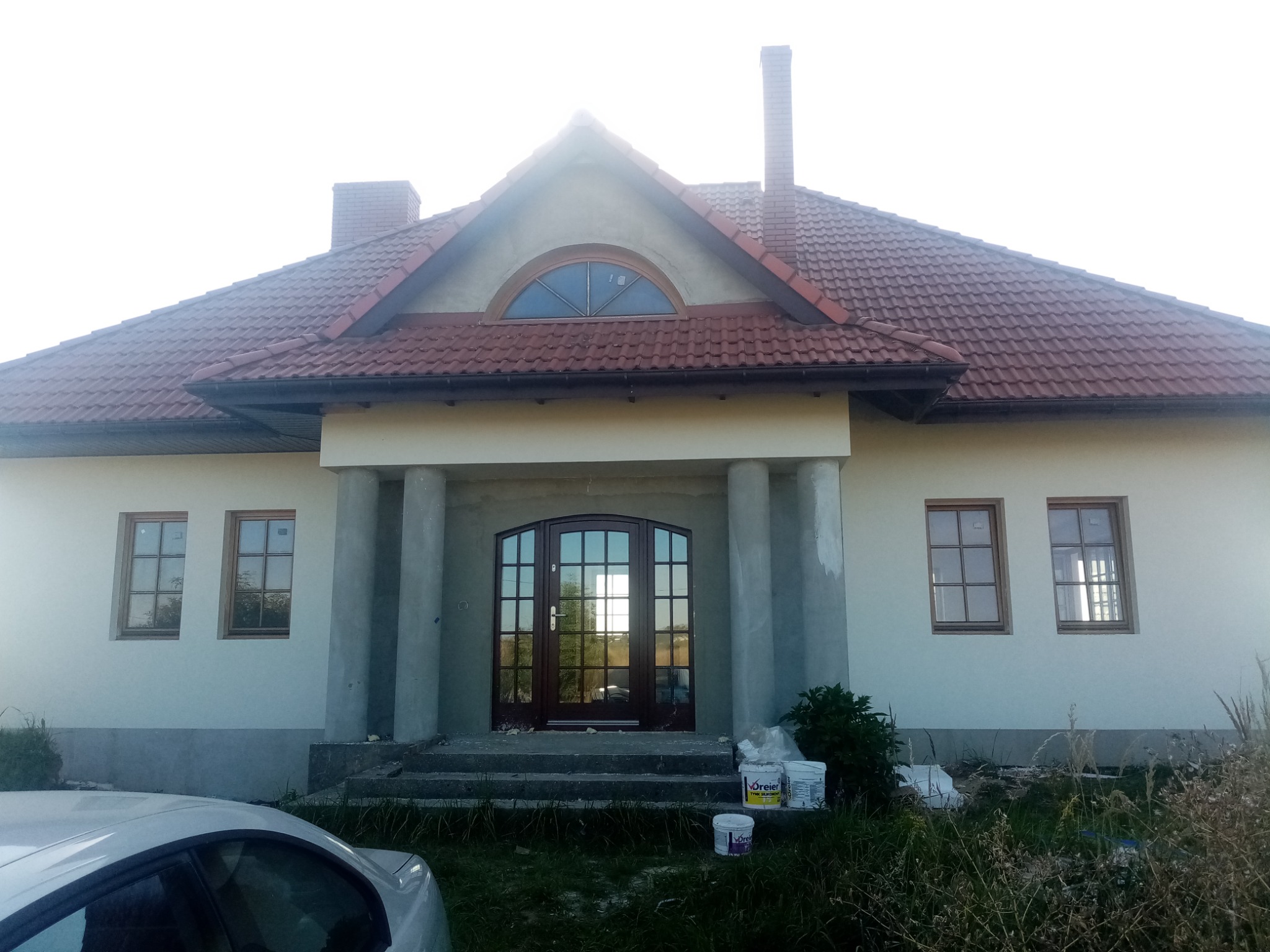 Dom jednorodzinny z kolumnami przed wejściem, widoczne ślady prac wykończeniowych na elewacji, farby w wiadrach przed wejściem, okna z drewnianymi ramami i czerwona dachówka.