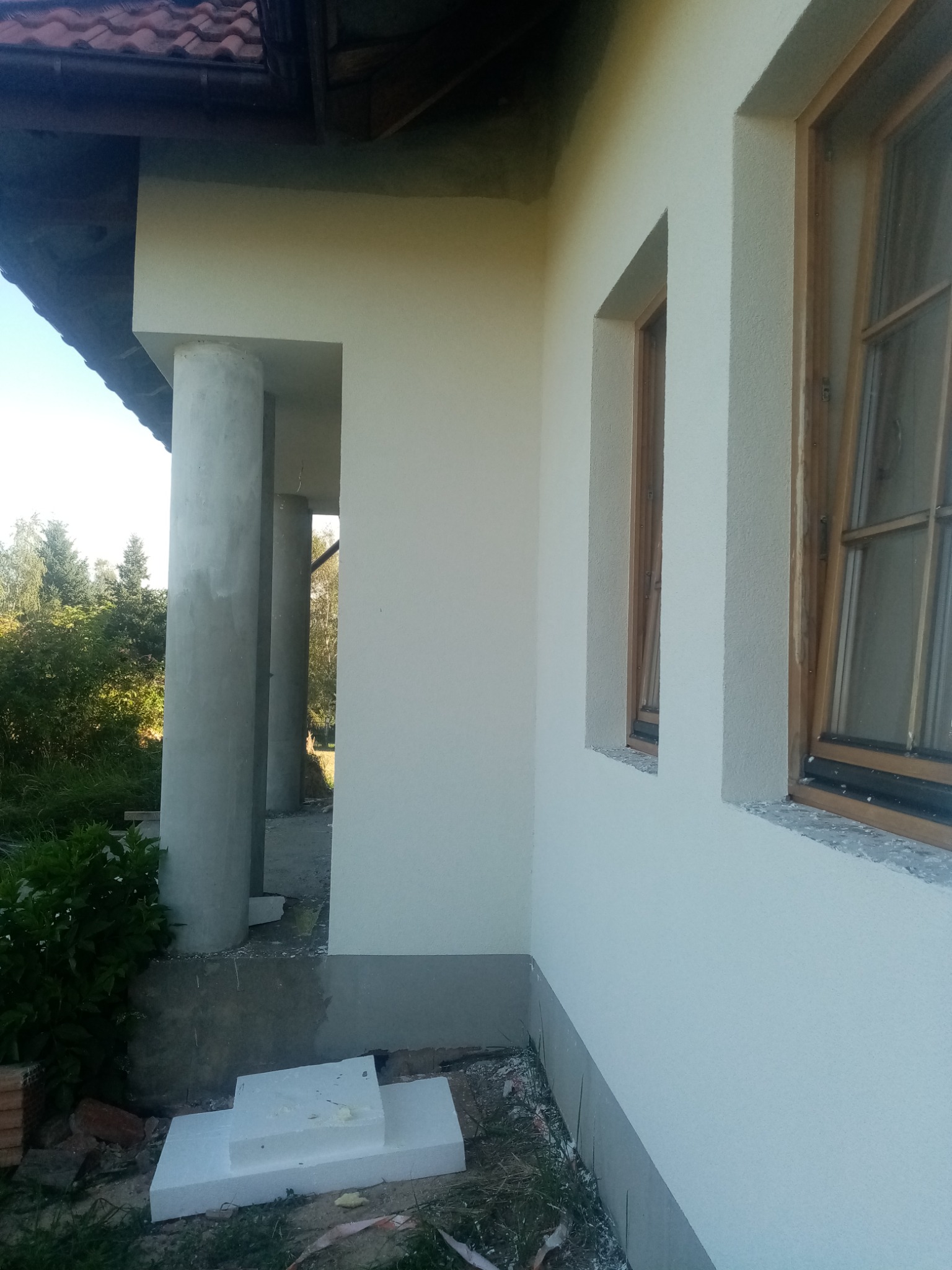 Fragment elewacji domu z oknami w drewnianych ramach i kolumnami podtrzymującymi zadaszenie, widoczne ślady prac wykończeniowych i styropianowe płyty.