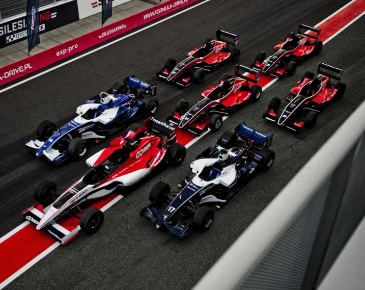Formuły wyścigowe ustawione na starcie, z widocznymi detalami malowania i numerami startowymi, na tle toru wyścigowego z reklamami sponsorów.