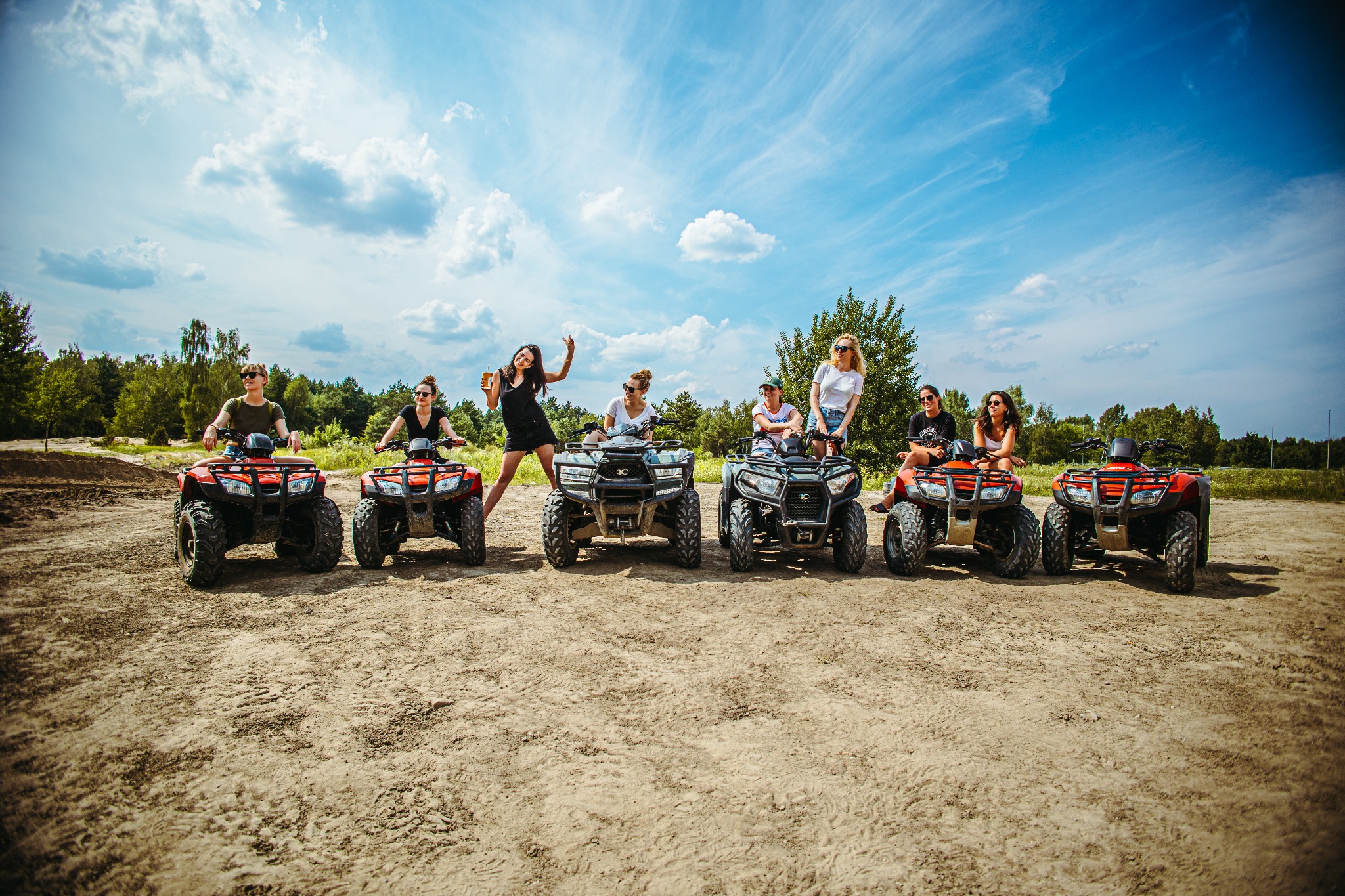 Grupa siedmiu osób pozuje na quadach na piaszczystym terenie pod błękitnym niebem z chmurami. Jedna osoba stoi obok quada z podniesioną ręką i napojem.