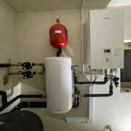 Zamontowany, biały piec gazowy Panasonic Aquarea z podłączonym zasobnikiem Galmet, widoczny czerwony zbiornik wyrównawczy i orurowanie, zainstalowane w pomieszczeniu z oknem.