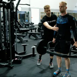 Trener personalny z Krakowa asekuruje klienta podczas podnoszenia ciężaru na trap barze w siłowni, w tle widoczne inne osoby ćwiczące i sprzęt do ćwiczeń.