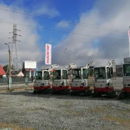 Rząd nowych, fabrycznie czystych minikoparek marki Takeuchi TD225 ustawionych na żwirowym placu przed budynkiem dealera w słoneczny dzień.