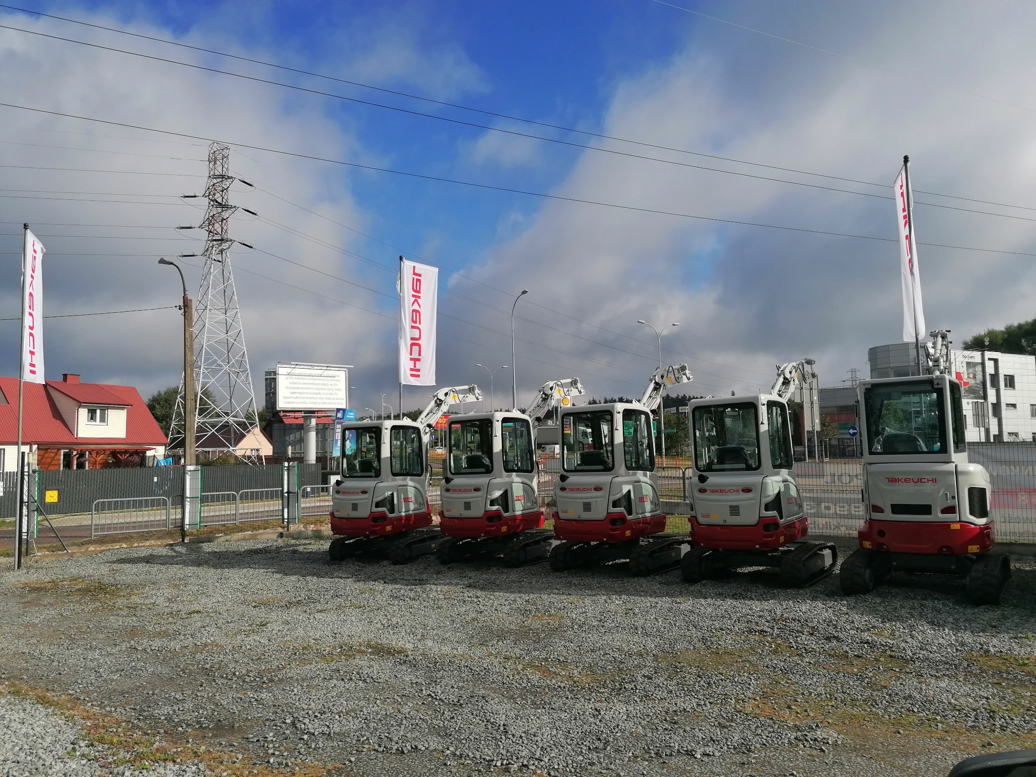 Rząd nowych, fabrycznie czystych minikoparek marki Takeuchi TD225 ustawionych na żwirowym placu przed budynkiem dealera w słoneczny dzień.
