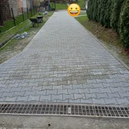 Długa, szara ścieżka z kostki brukowej w kształcie litery I, zakończona metalową kratką odwadniającą, prowadząca w kierunku domu z zielonym trawnikiem i wysokimi tujami po prawej stronie, widoczne...