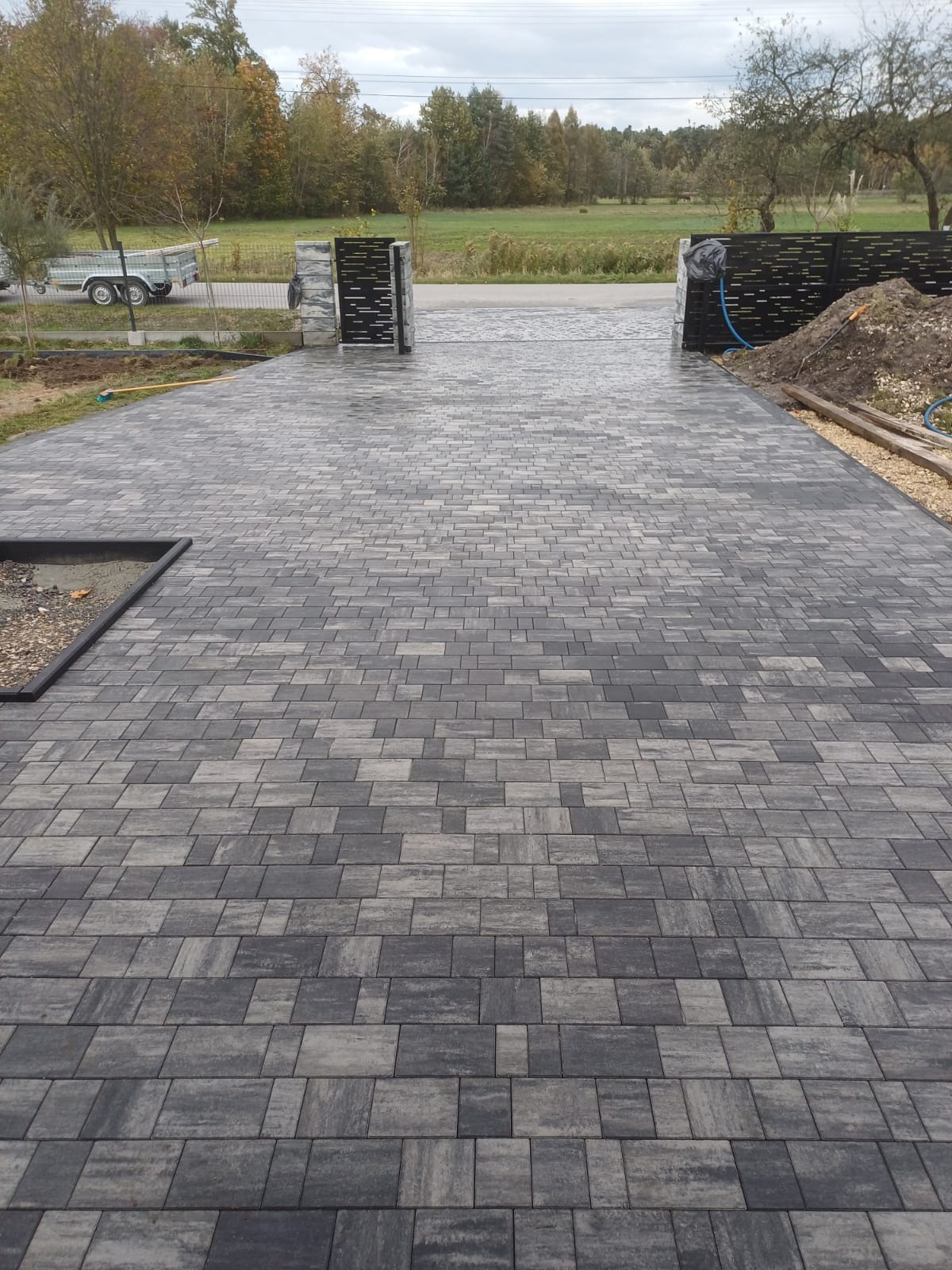 Ułożona szara kostka brukowa prowadząca do metalowej bramy wjazdowej, widoczny fragment ogrodu i przyczepka samochodowa w tle.