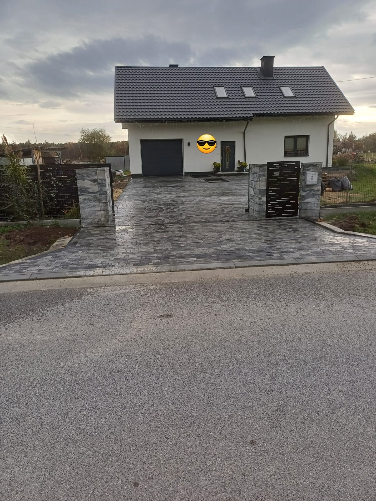 Ułożona, szara kostka brukowa przed nowoczesnym domem z szarą dachówką i garażem, widoczny fragment ogrodzenia z metalowymi elementami i kamiennymi słupkami.