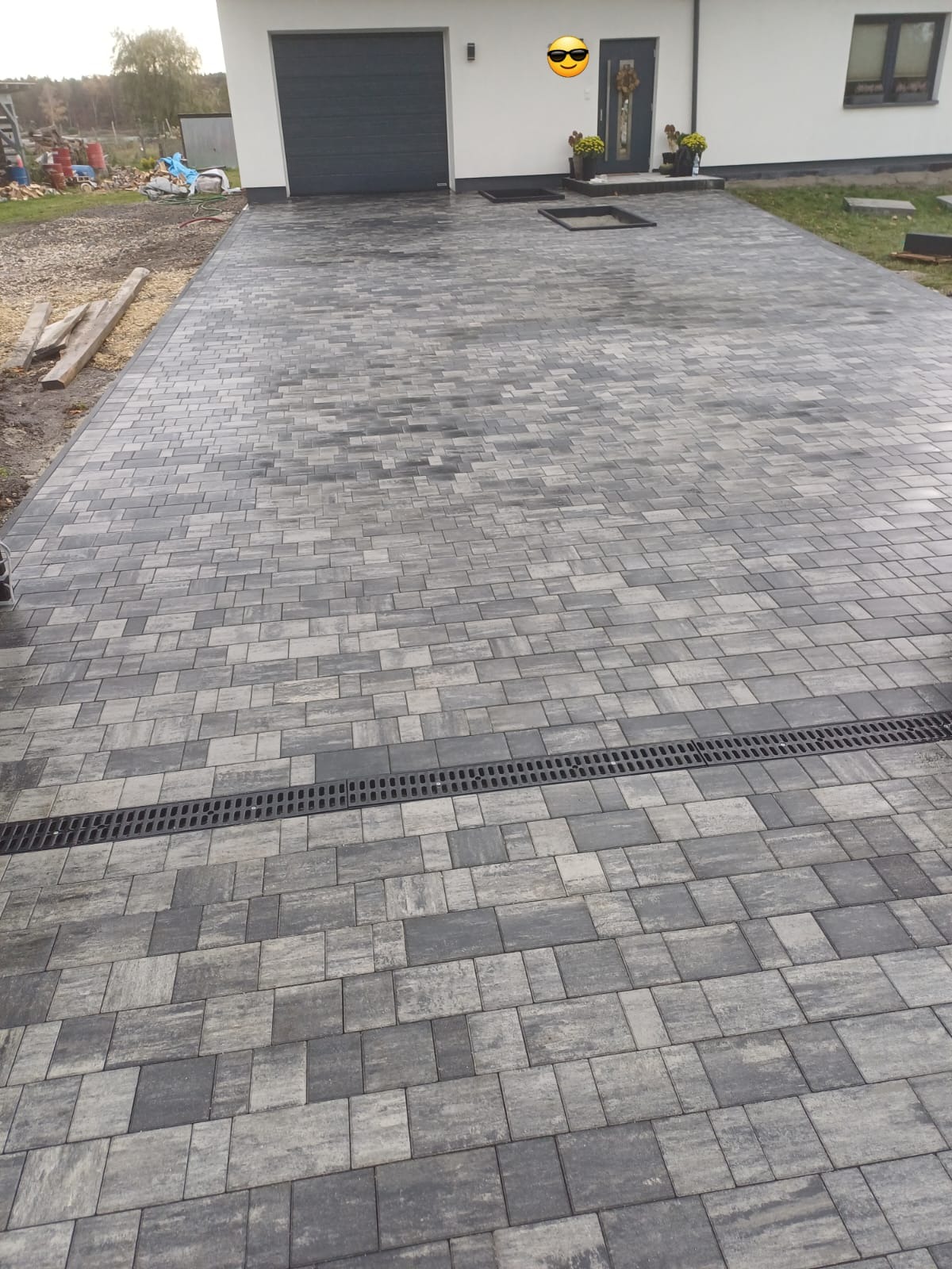 Ułożona kostka brukowa w odcieniach szarości z odpływem liniowym przed domem z bramą garażową.