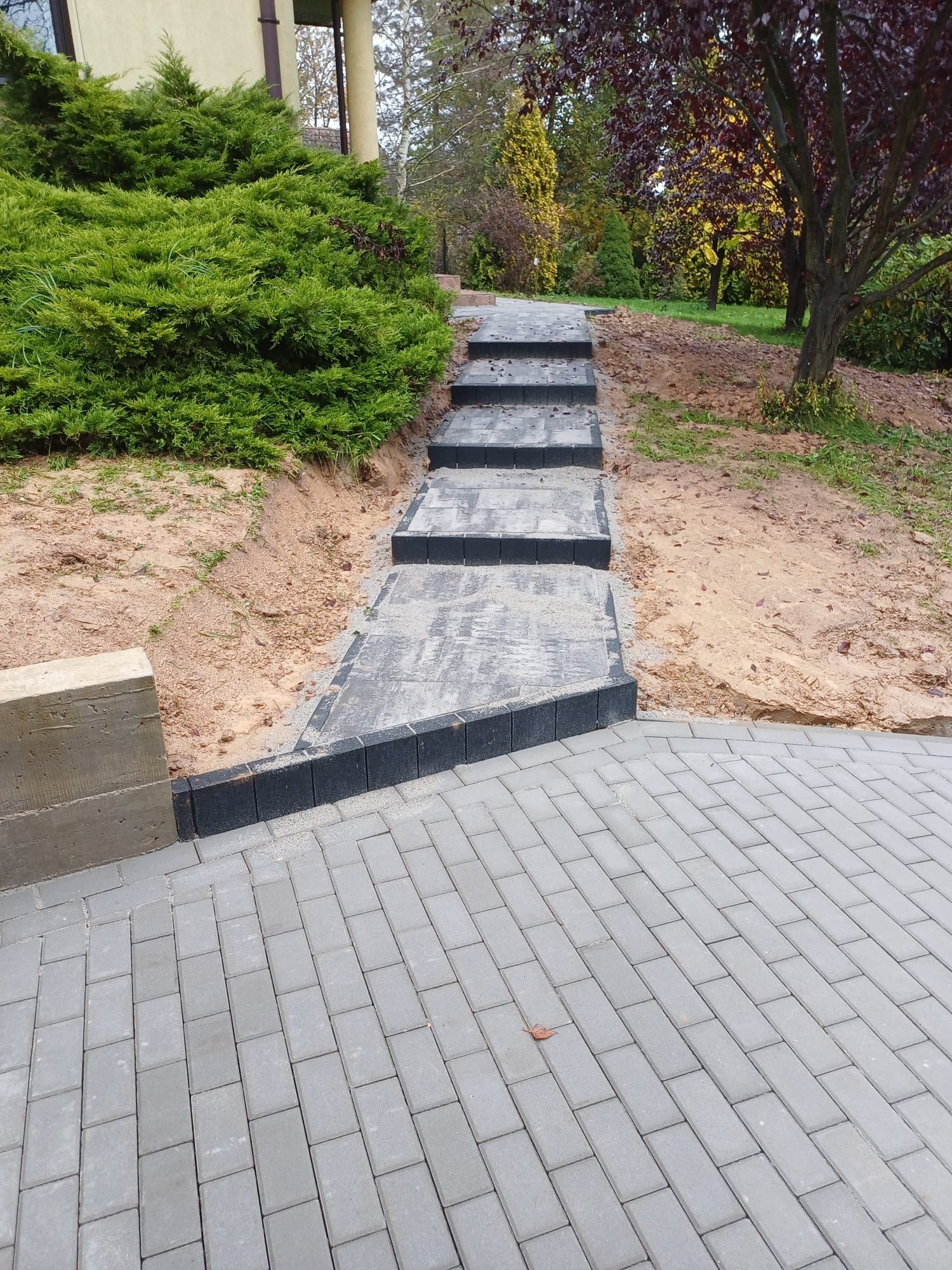 Ścieżka z szarych prostokątnych kostek brukowych prowadząca do schodów z ciemnych kostek, otoczona zielenią i ziemią.
