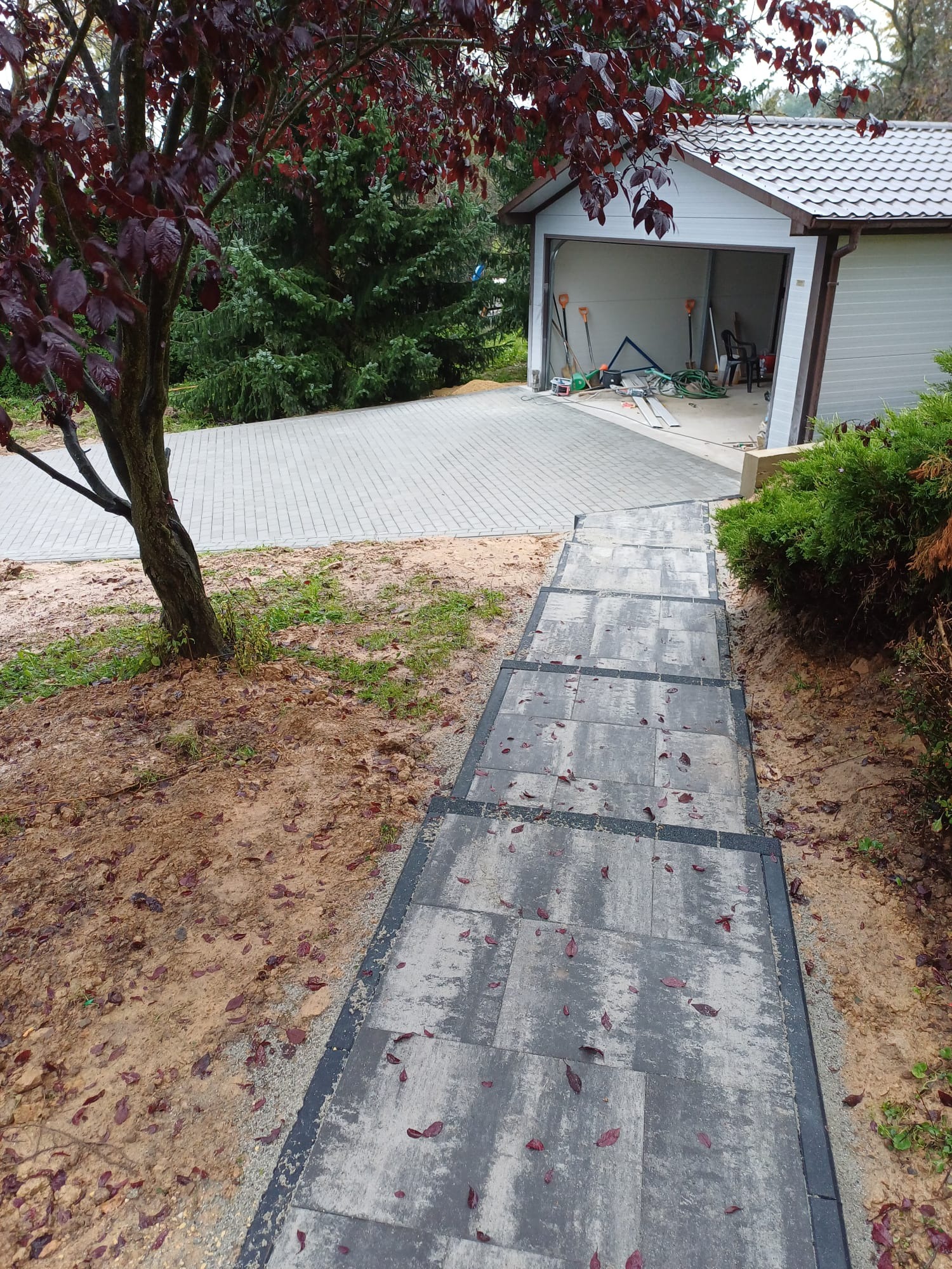 Schody z szarej kostki brukowej z ciemnymi obrzeżami prowadzące do podjazdu z kostki i garażu z narzędziami w tle, częściowo przysypane opadłymi liśćmi