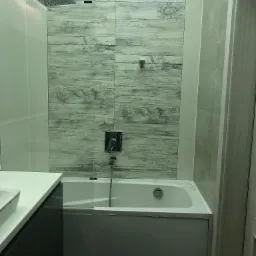 Wykończona łazienka w Węgrzcach Wielkich z wanną obudowaną szarą zabudową, nad którą zamontowano szklany panel i płytki imitujące drewno z motywem kwiatowym, oraz deszczownicę.