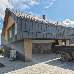 Fliśnik Zakład Remontowo Budowlany - Nowoczesny dom z grafitową elewacją panelową i drewnianymi akcentami, widok z zewnątrz, z fragmentem ciężarówki na podjeździe i pracami wykończeniowymi w tle.
