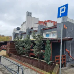 Budynek w trakcie ocieplania elewacji, widoczne płyty styropianowe, rusztowania i instalacja odwadniająca. Na pierwszym planie zadbane drzewka iglaste i znak drogowy.