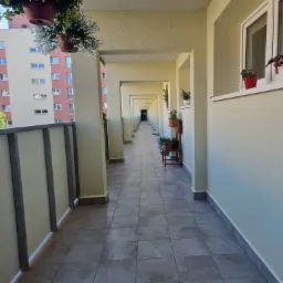 Długi, zadaszony balkon w bloku, wyłożony szarą terakotą, z widokiem na kolejne balkony i odległy budynek mieszkalny, dekorowany wiszącymi i stojącymi doniczkami z kwiatami.