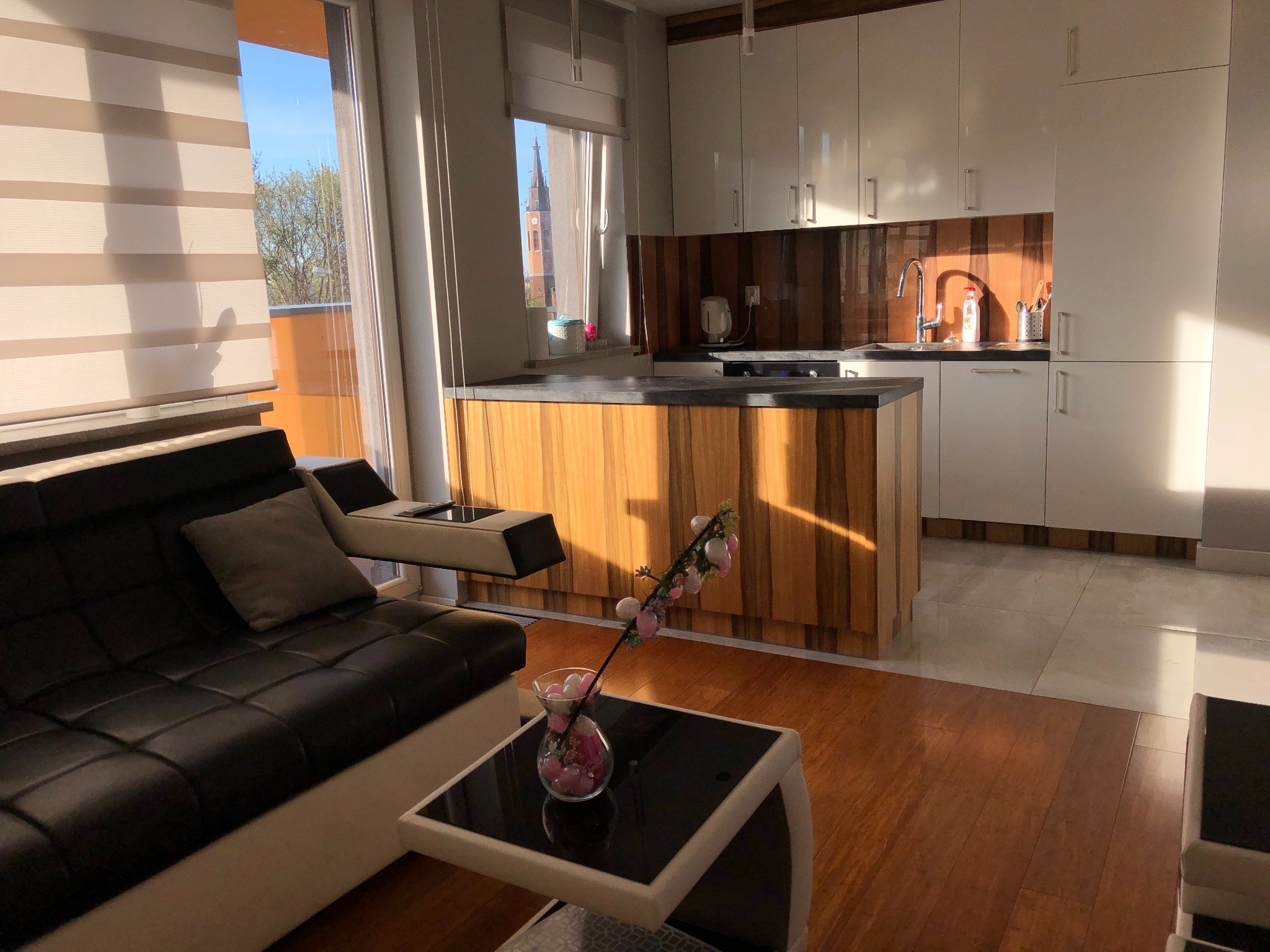 Nowoczesne wnętrze mieszkania z aneksem kuchennym, widok na zabudowę kuchenną z drewnianymi akcentami i wyspą, w tle kościół przez okno, w salonie czarna skórzana sofa i stolik kawowy z dekoracją.