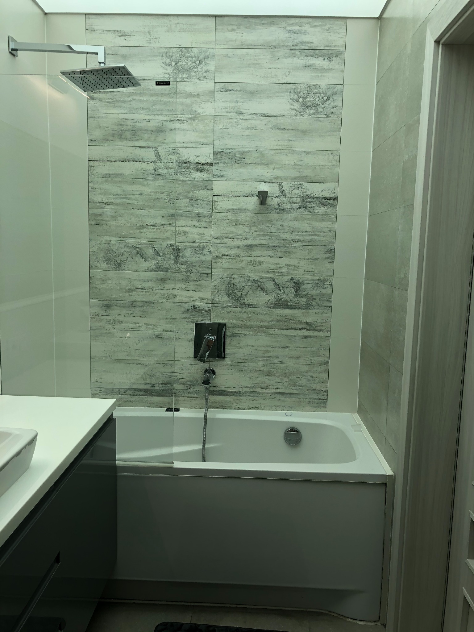 Wykończona łazienka w Węgrzcach Wielkich z wanną obudowaną szarą zabudową, nad którą zamontowano szklany panel i płytki imitujące drewno z motywem kwiatowym, oraz deszczownicę.