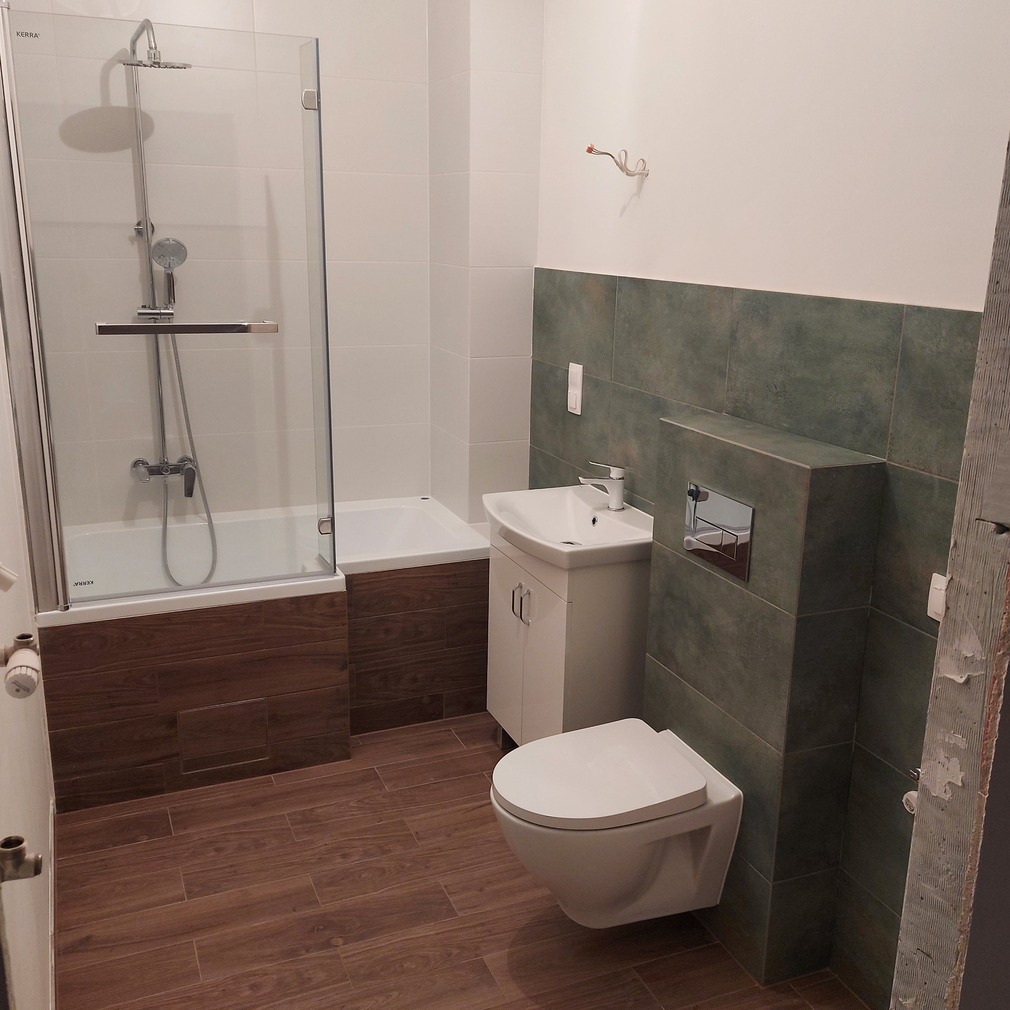 Nowoczesna łazienka z kabiną prysznicową, umywalką z szafką i podwieszaną toaletą; ściany wykończone białymi i zielonymi płytkami, podłoga imitująca drewno.