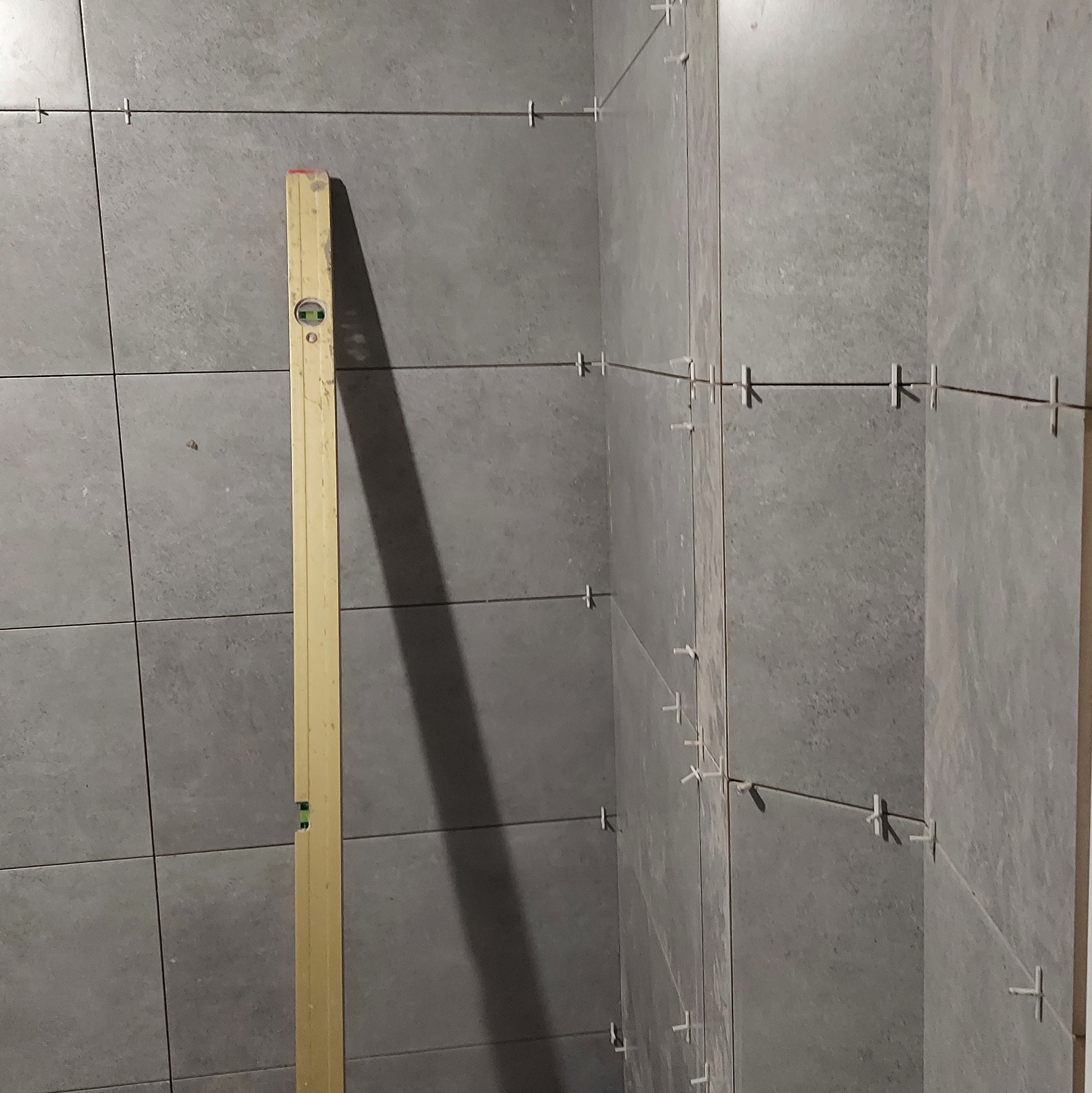 Układanie szarych płytek w rogu pomieszczenia, poziomica przy ścianie, krzyżyki dystansowe w fugach.