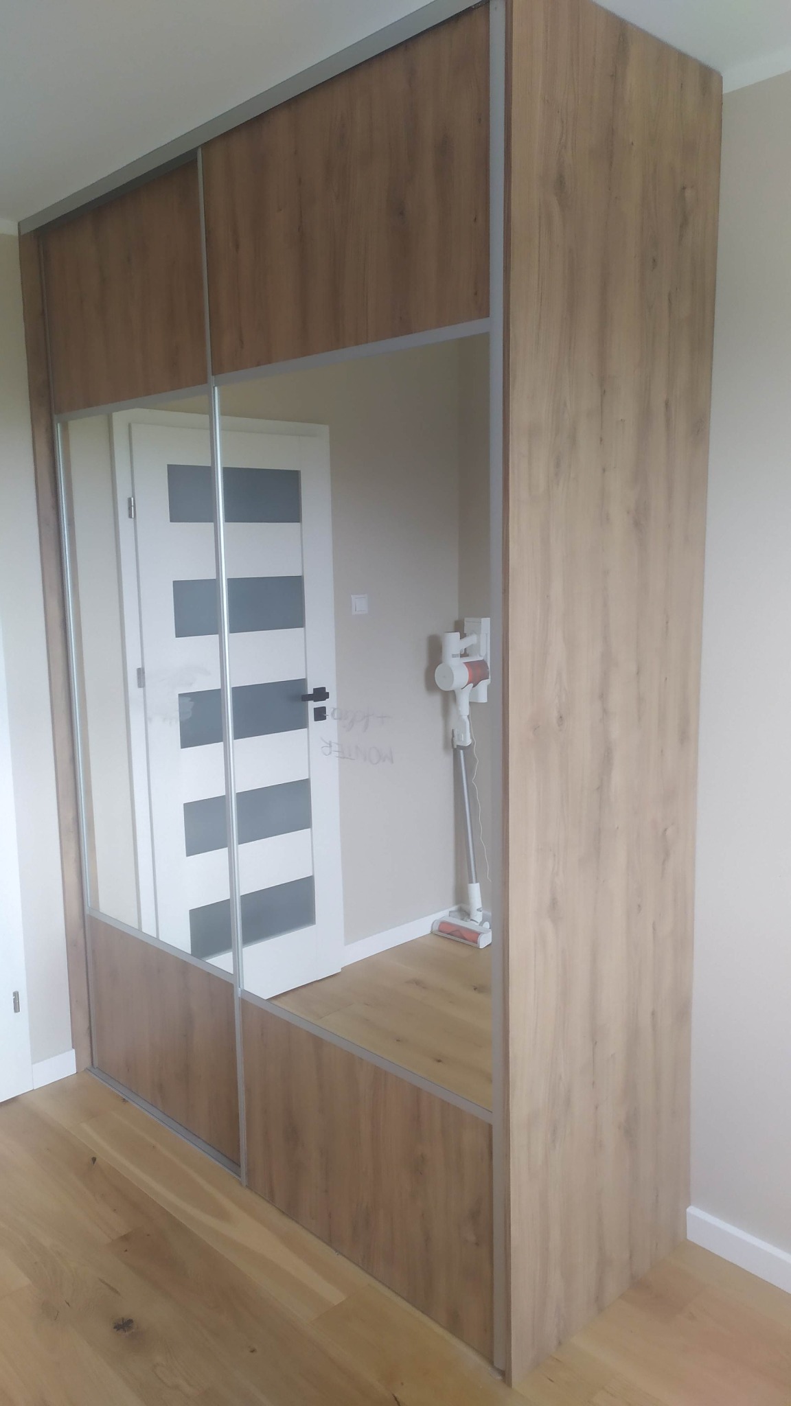 Szafa wnękowa z przesuwnymi lustrzanymi drzwiami w kolorze drewna, częściowo odbijająca białe drzwi z szarymi panelami i odkurzacz pionowy.