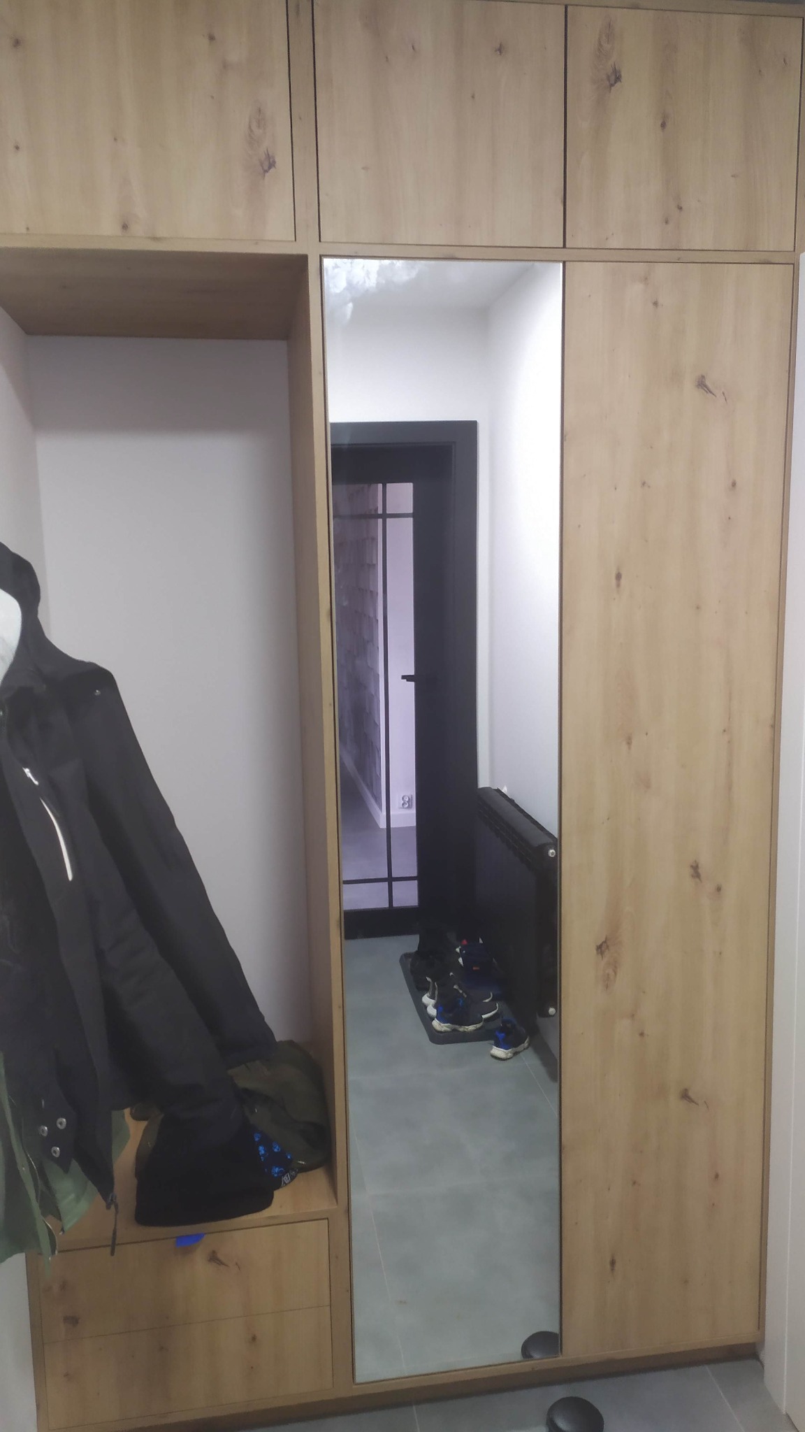 Drewniana szafa z lustrem w korytarzu, widoczne ubrania na wieszaku i odbicie butów na wycieraczce.