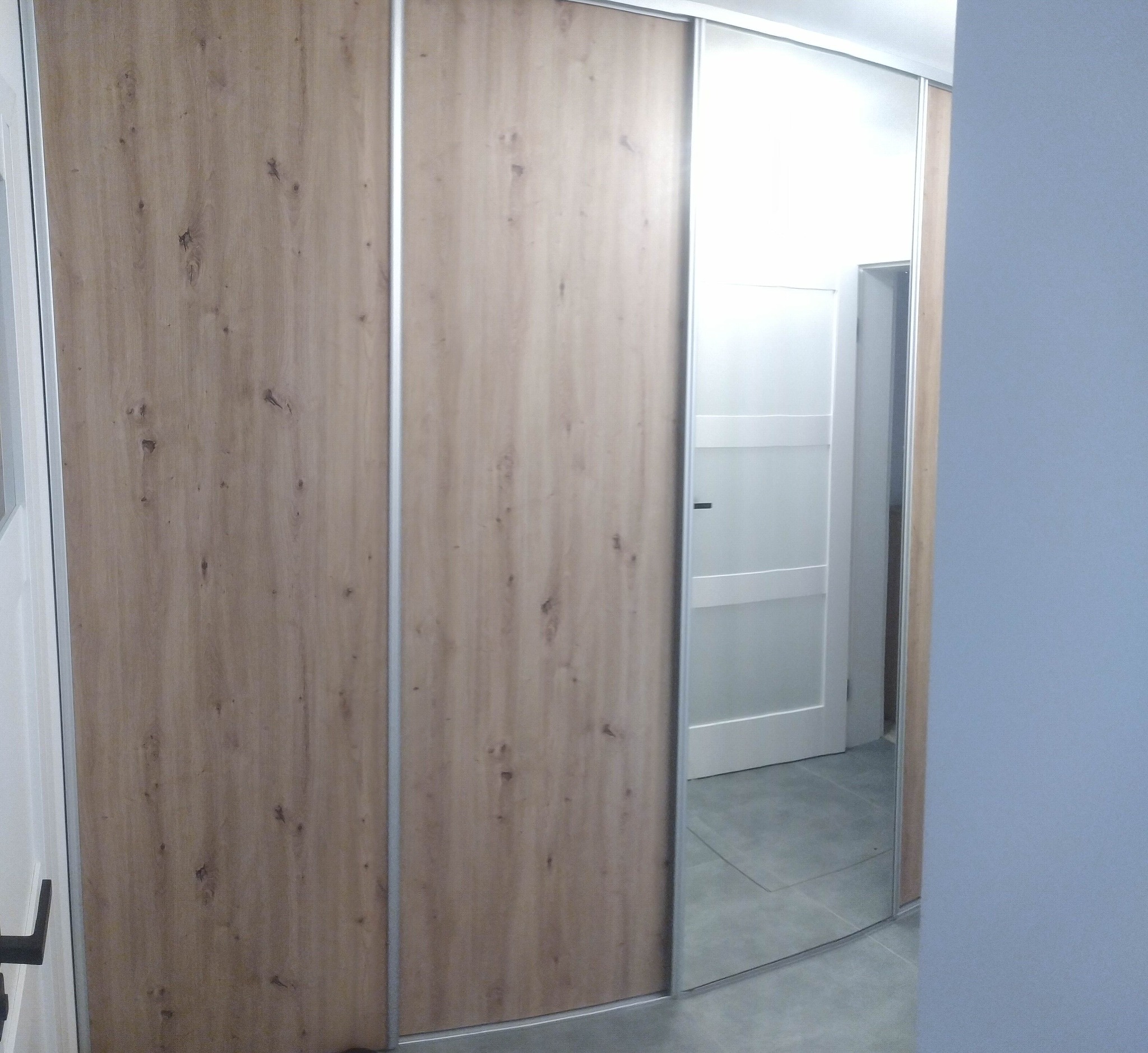 Zamknięta szafa wnękowa z przesuwnymi drzwiami: dwoje drzwi z płyty drewnopodobnej w odcieniach beżu i jedno lustrzane, srebrne obramowanie.