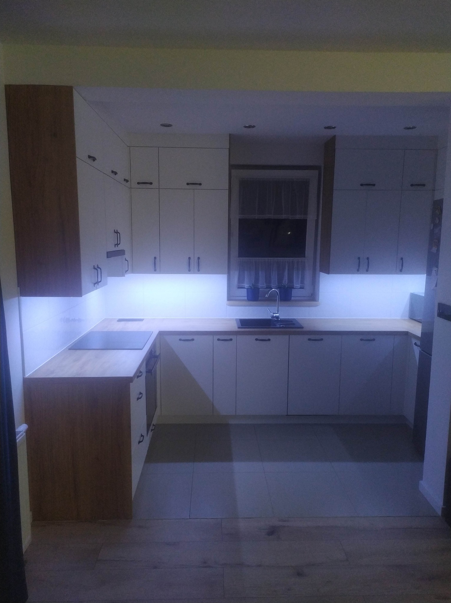 Nowoczesna kuchnia z białymi szafkami i drewnianymi blatami, oświetlona subtelnym światłem LED pod szafkami wiszącymi.