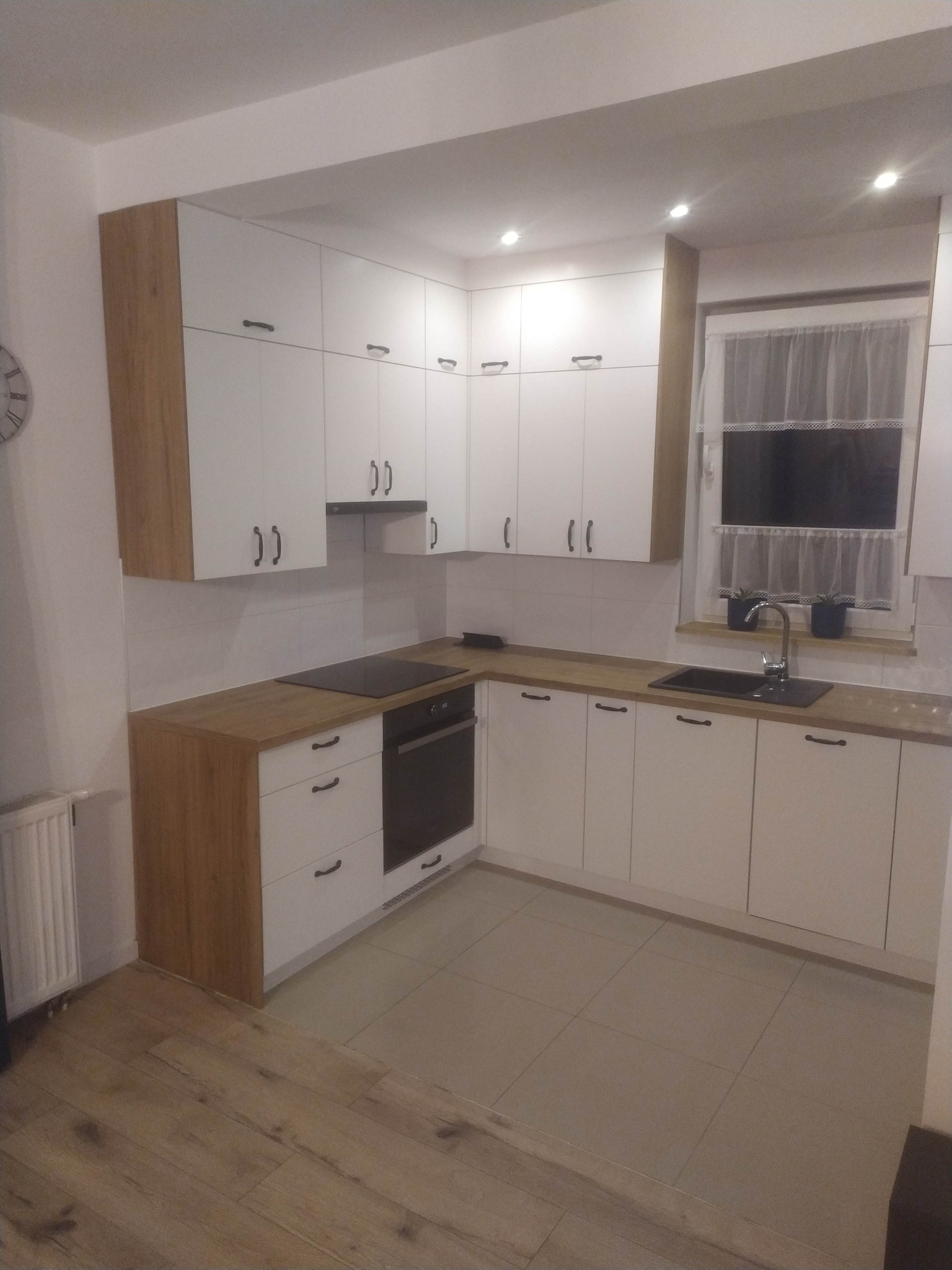 Jasna kuchnia w kształcie litery L z białymi frontami i drewnianymi blatami, czarne uchwyty, płyta indukcyjna i piekarnik, okno z firanką i doniczkami.