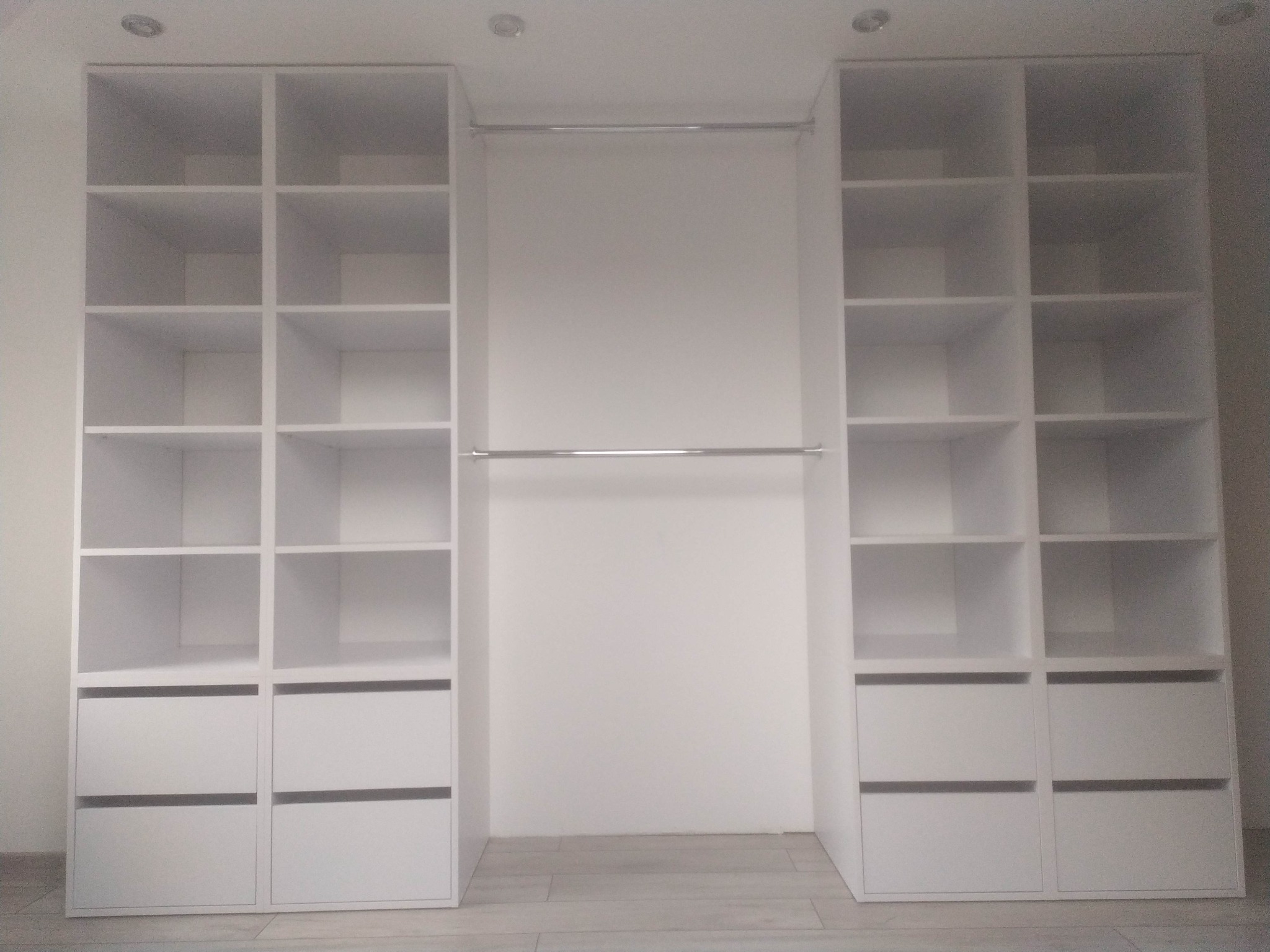 Biała zabudowa wnęki z półkami i szufladami po bokach, drążek na ubrania w środkowej części.