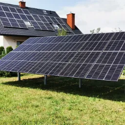 Panele słoneczne zamontowane na gruncie przed domem z panelami na dachu. Widoczne metalowe podpory paneli i trawnik.