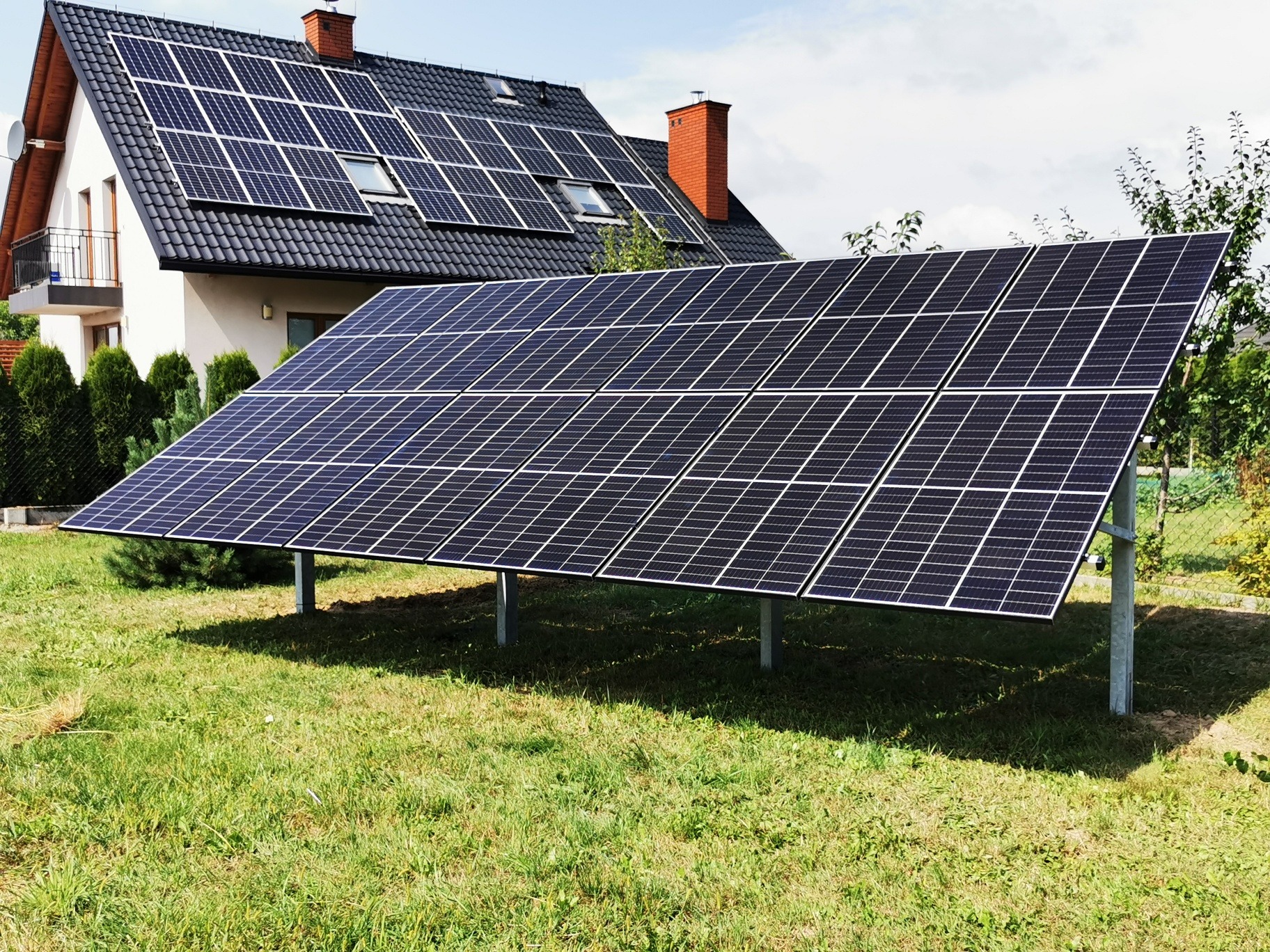 Panele słoneczne zamontowane na gruncie przed domem z panelami na dachu. Widoczne metalowe podpory paneli i trawnik.