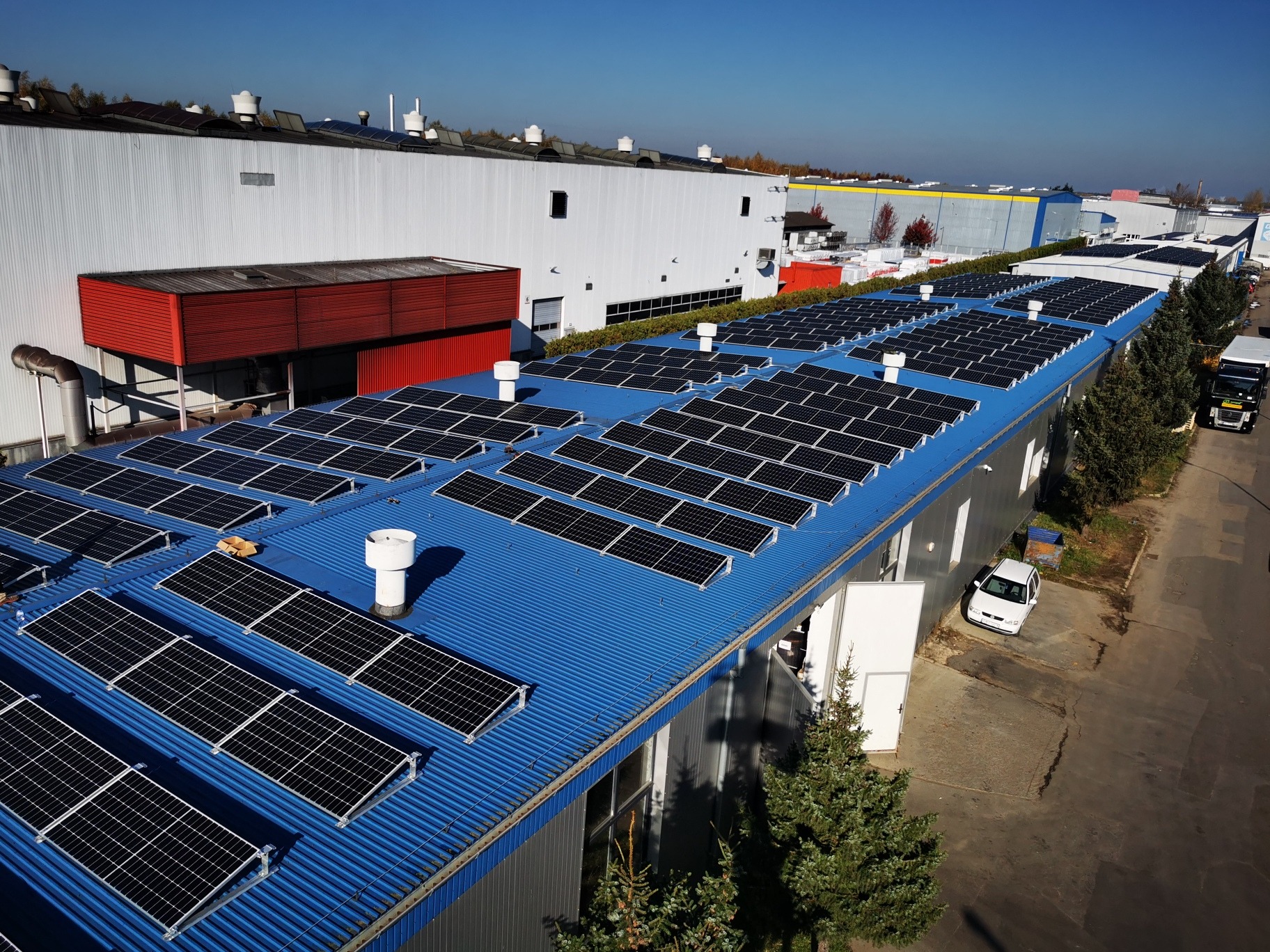 Widok z góry na dużą instalację fotowoltaiczną na niebieskim dachu hali przemysłowej, widoczne kominki wentylacyjne i fragmenty innych budynków w tle.