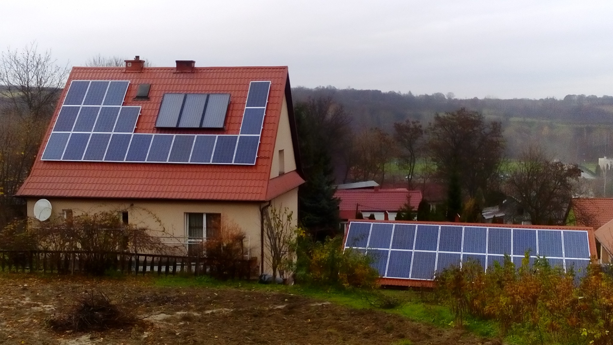 Dom jednorodzinny z panelami słonecznymi na dachu z czerwonej dachówki, dodatkowe panele na dachu budynku gospodarczego w tle, pochmurny dzień.
