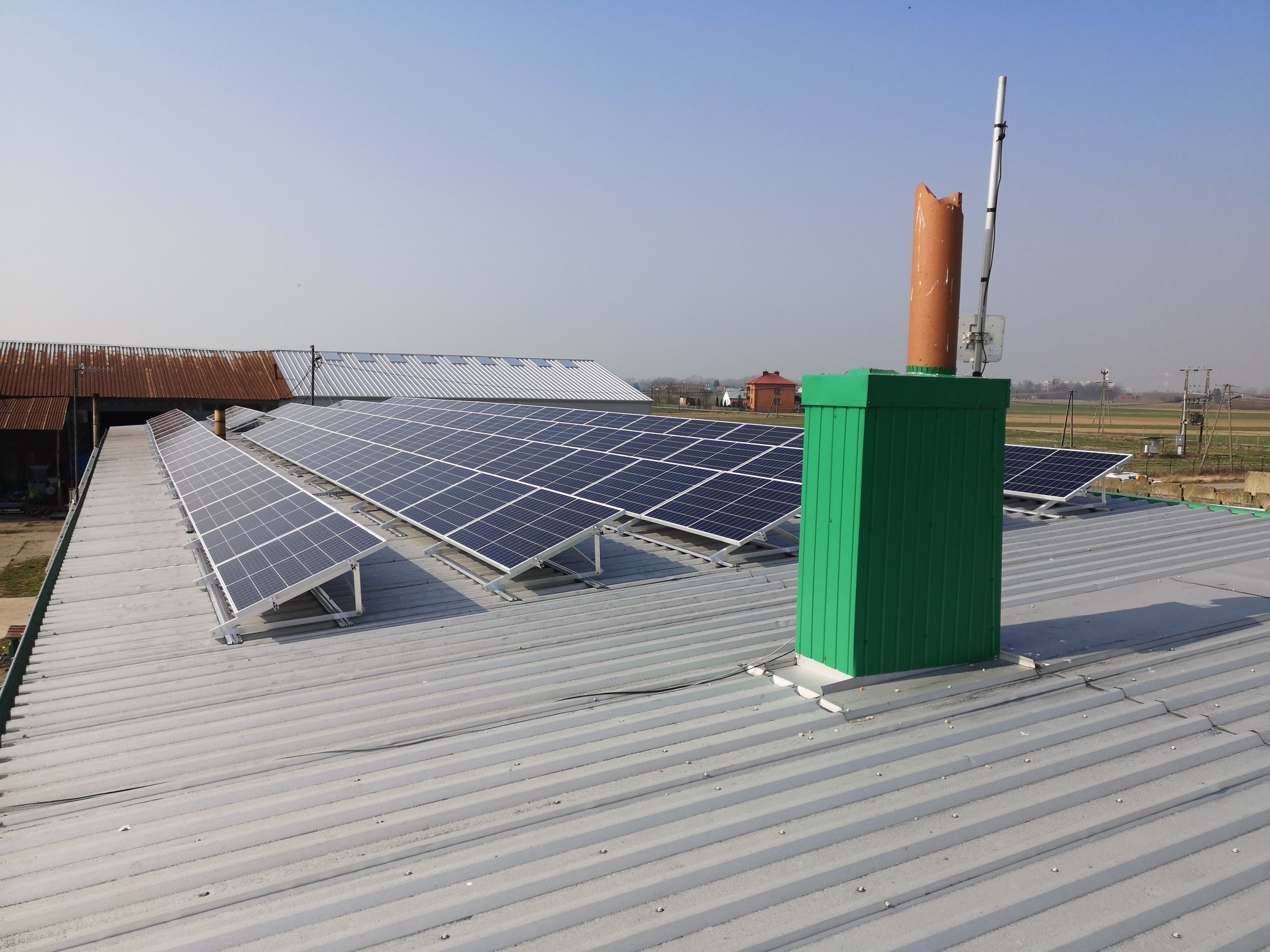 Panele słoneczne na dachu budynku gospodarczego, widoczne panele, komin wentylacyjny w kolorze zielonym i anteny.