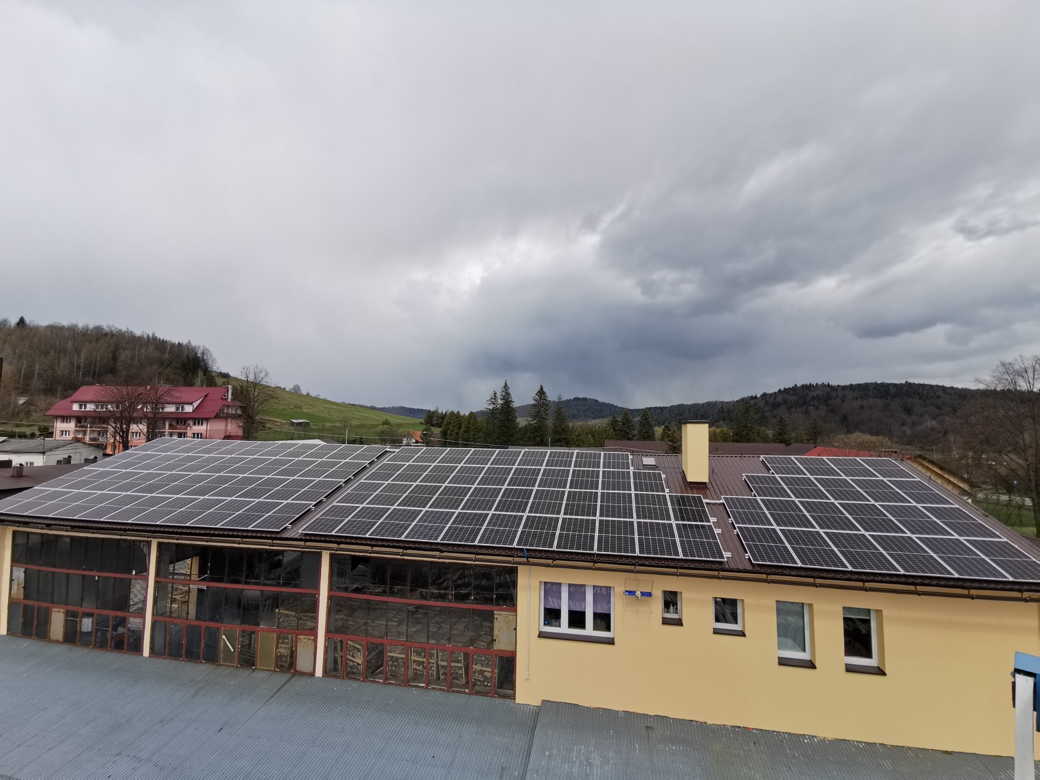 Instalacja paneli słonecznych na dachu budynku przemysłowego z widocznymi oknami i elementami konstrukcyjnymi, pochmurne niebo w tle.