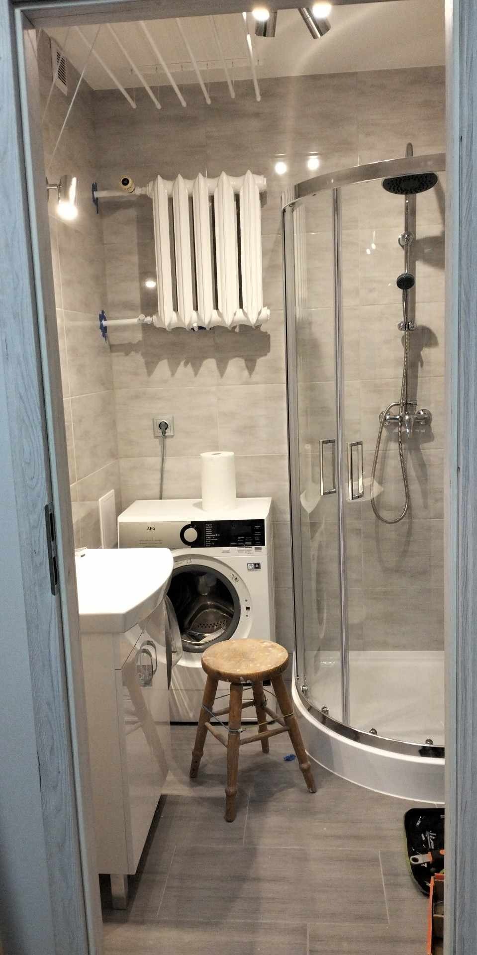 Mała łazienka po remoncie, z kabiną prysznicową, pralką AEG, grzejnikiem żeberkowym i drewnianym stołkiem.